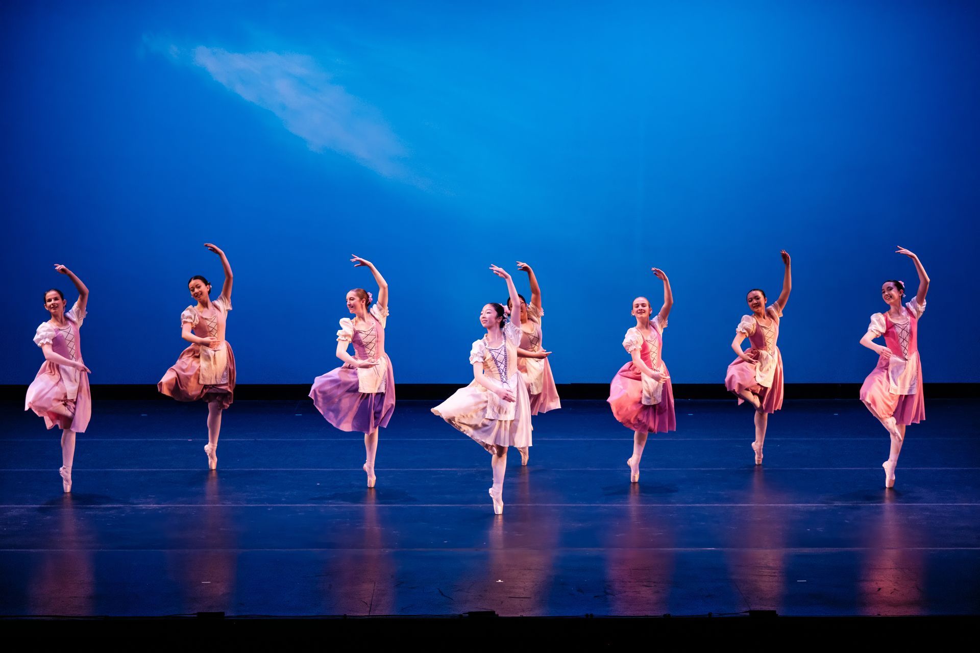 Seven ballet dancers in pink costumes performing synchronized poses on a blue-lit stage.