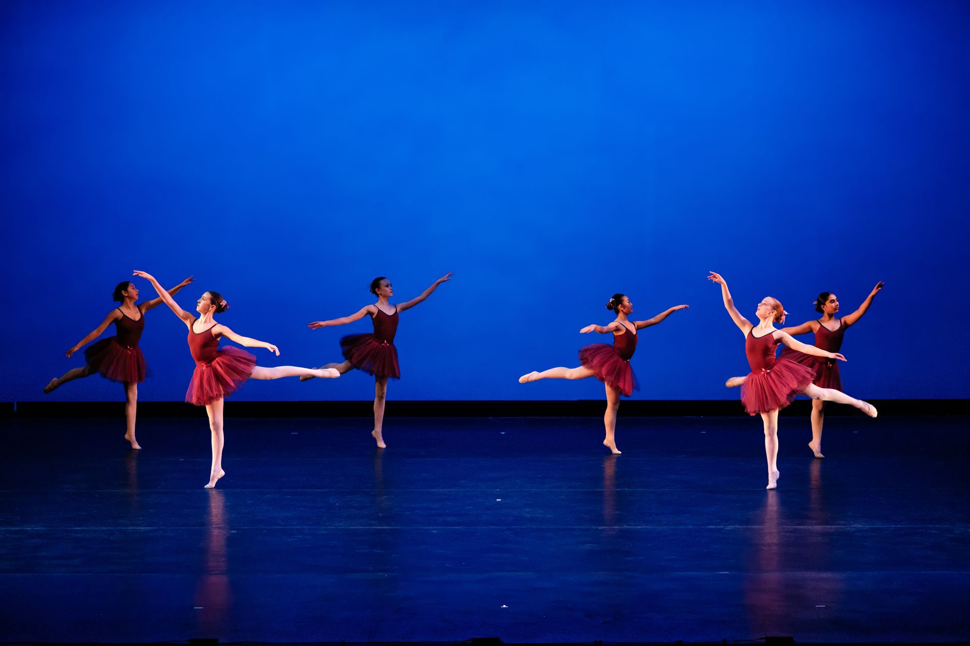Five dancers in red costumes perform ballet poses on a blue-lit stage.