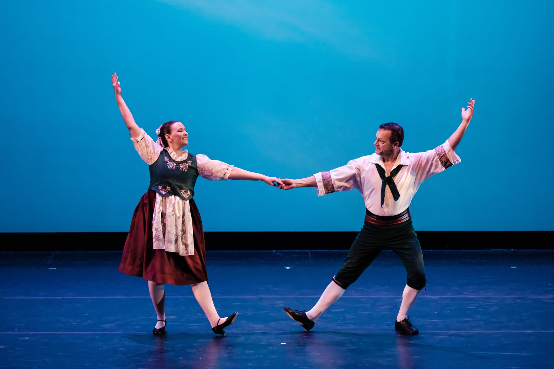 Two dancers holding hands on a blue-lit stage, arms raised in a lively pose