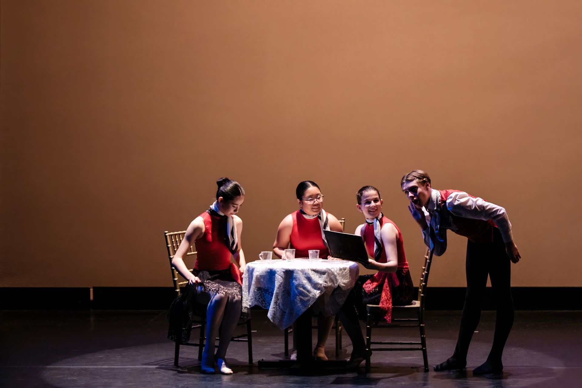 Four people sit and stand around a small table on a dim stage, lit by a warm spotlight.