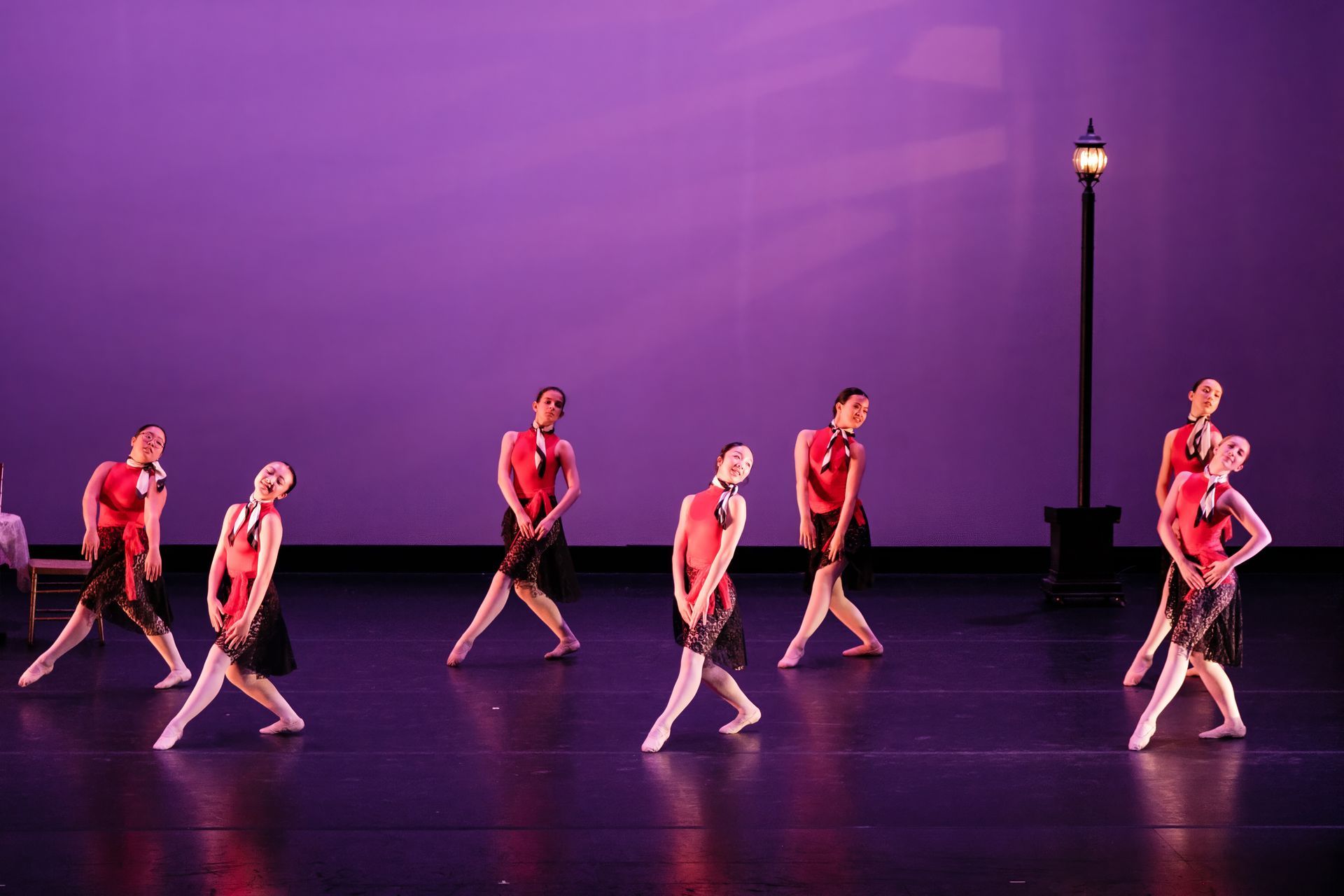 Dancers in red costumes perform on a stage with a purple backdrop and a lamppost prop