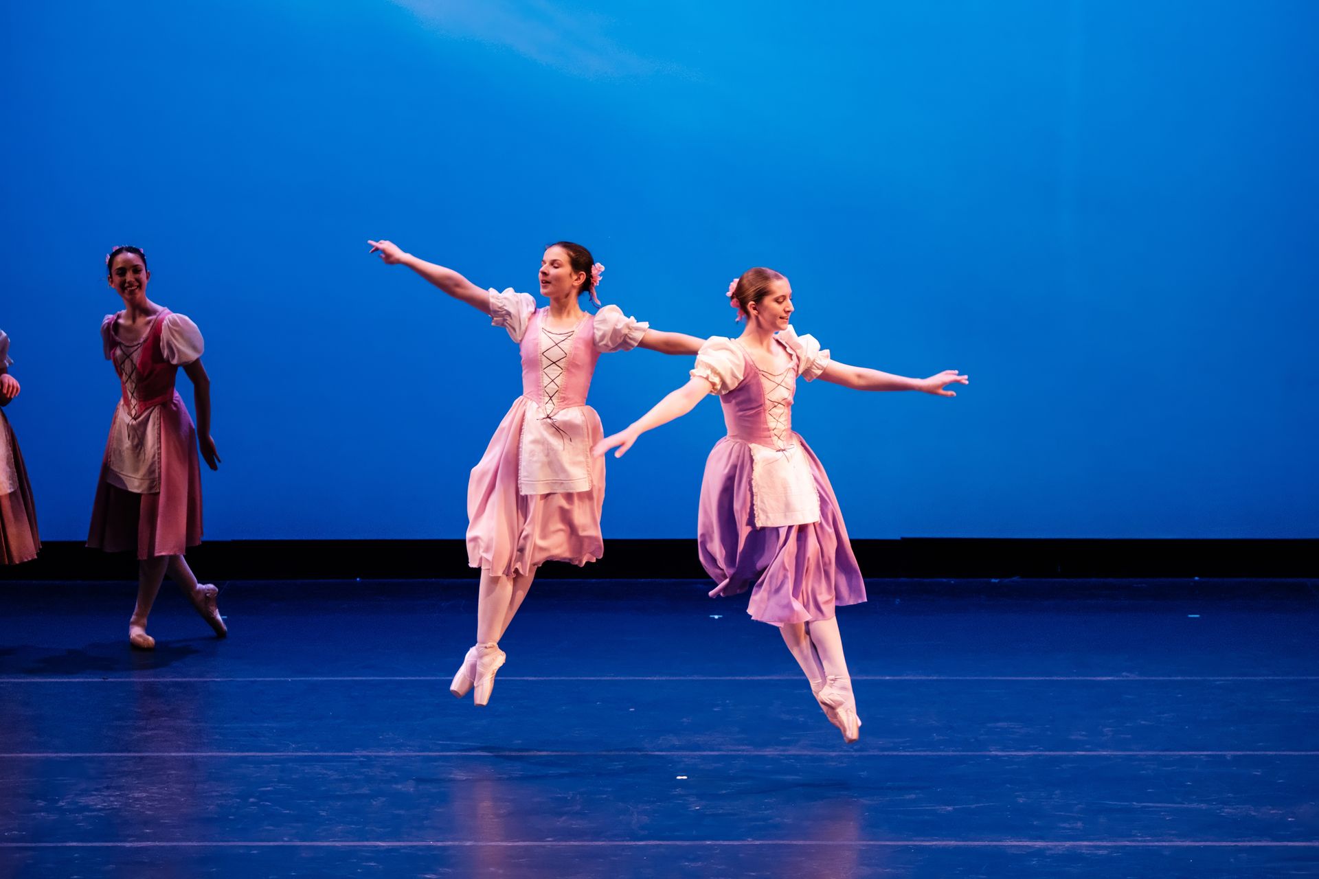 Two dancers in pink costumes leap across a blue-lit stage while another stands in the background