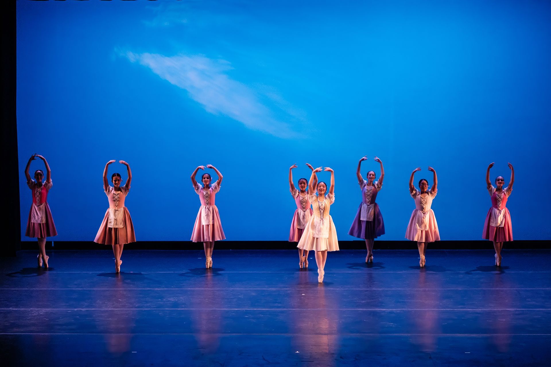 Dancers in colorful dresses perform on a blue-lit stage with arms raised overhead.