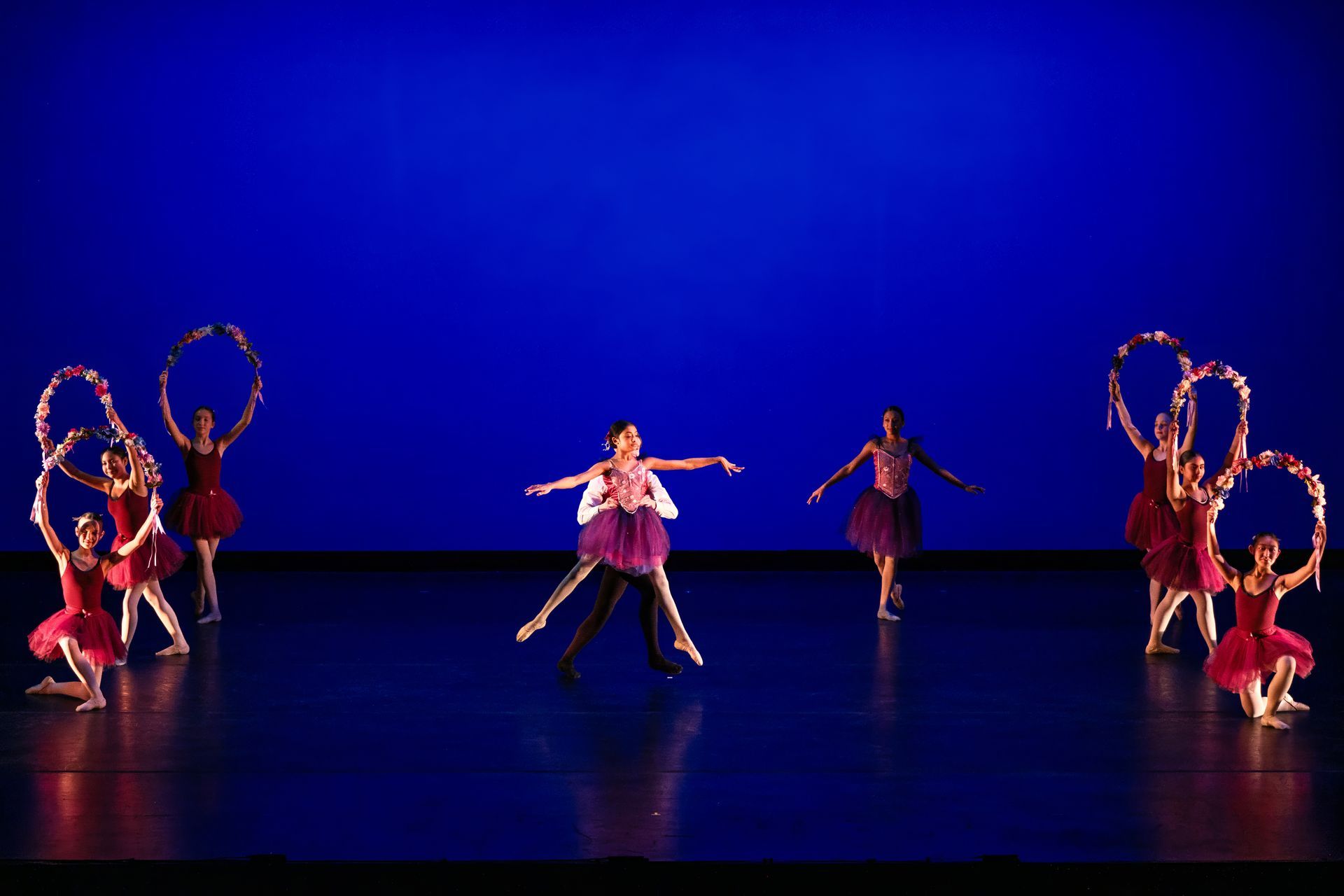 Dancers on a blue-lit stage pose with large circular props, centered around one performer in pink.
