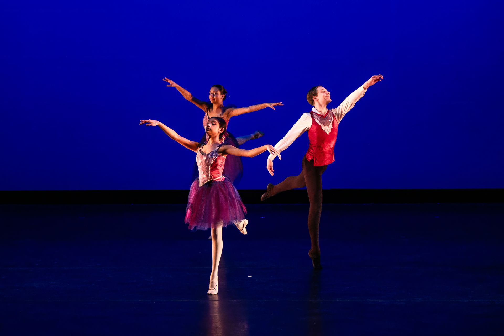 Three ballet dancers posing on a stage with a blue backdrop, one in a red costume in front.