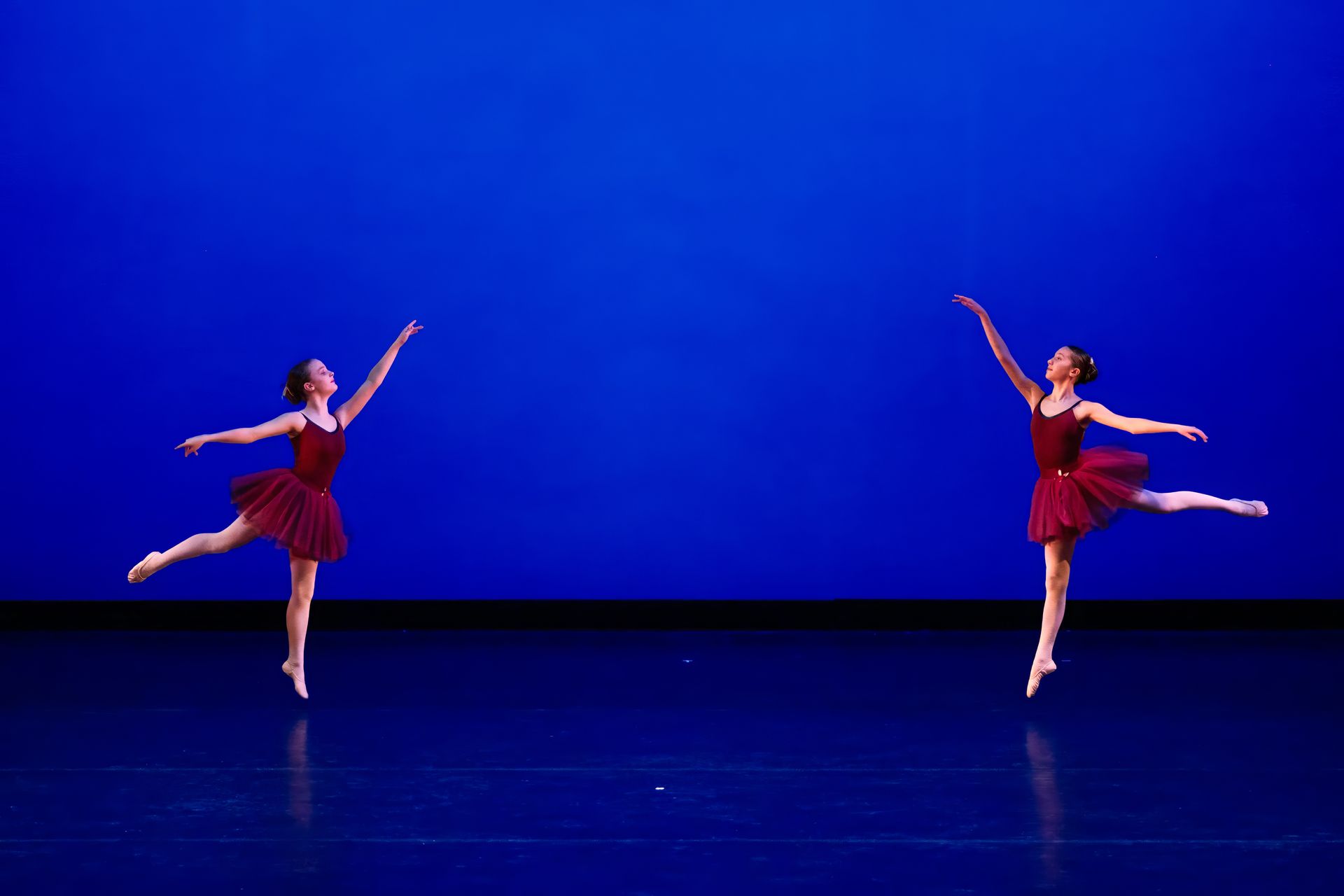 Two dancers in red costumes pose on a blue-lit stage, one balancing with a raised leg