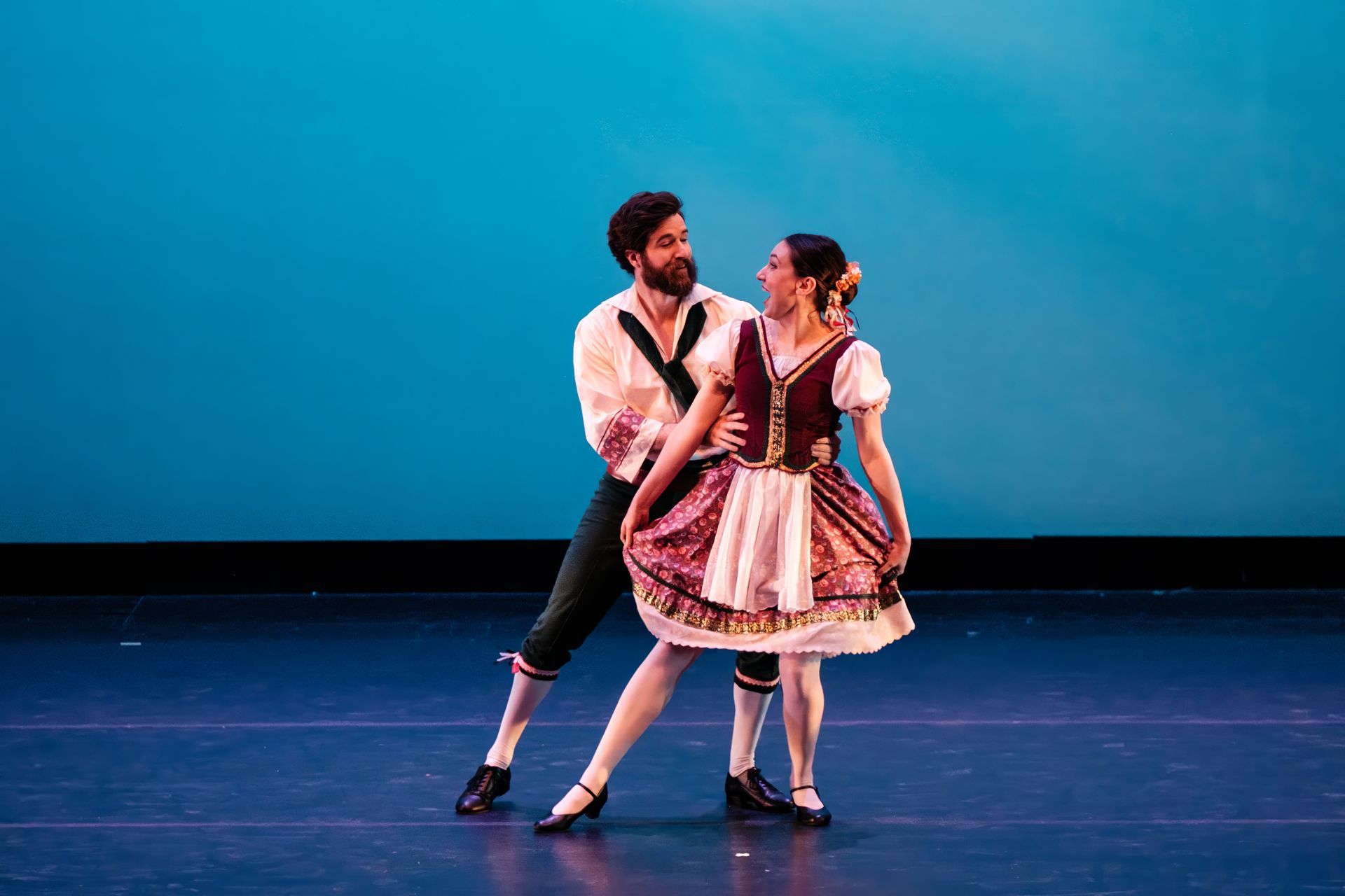 Two dancers in traditional costumes posing on a theater stage against a blue backdrop