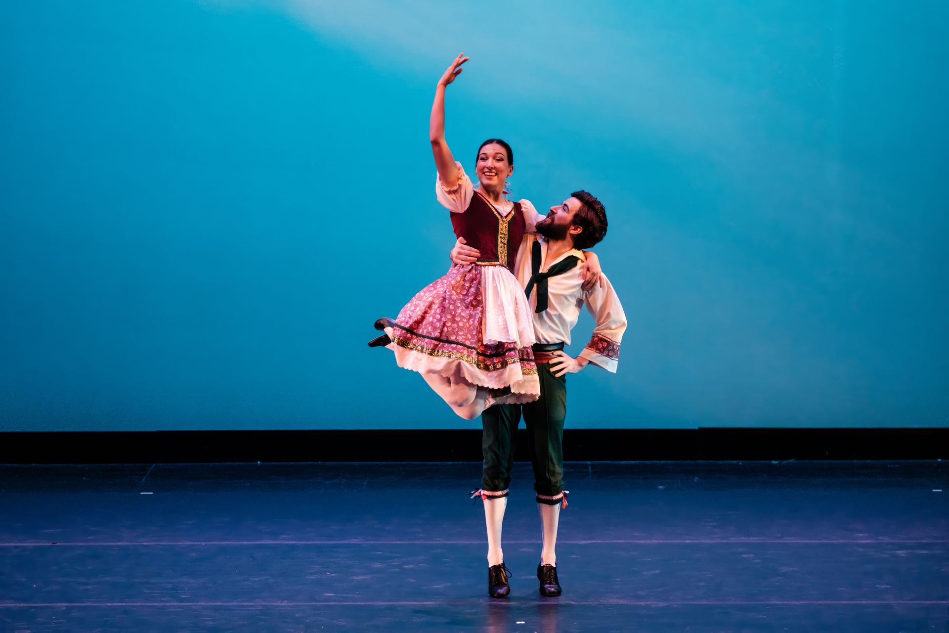 Dancer in pink tutu lifts a partner onstage against a blue backdrop