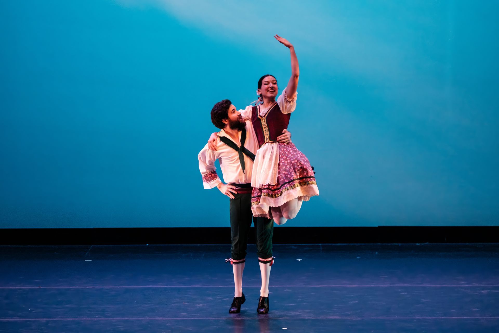 Two dancers on a stage, one lifting the other against a blue backdrop.