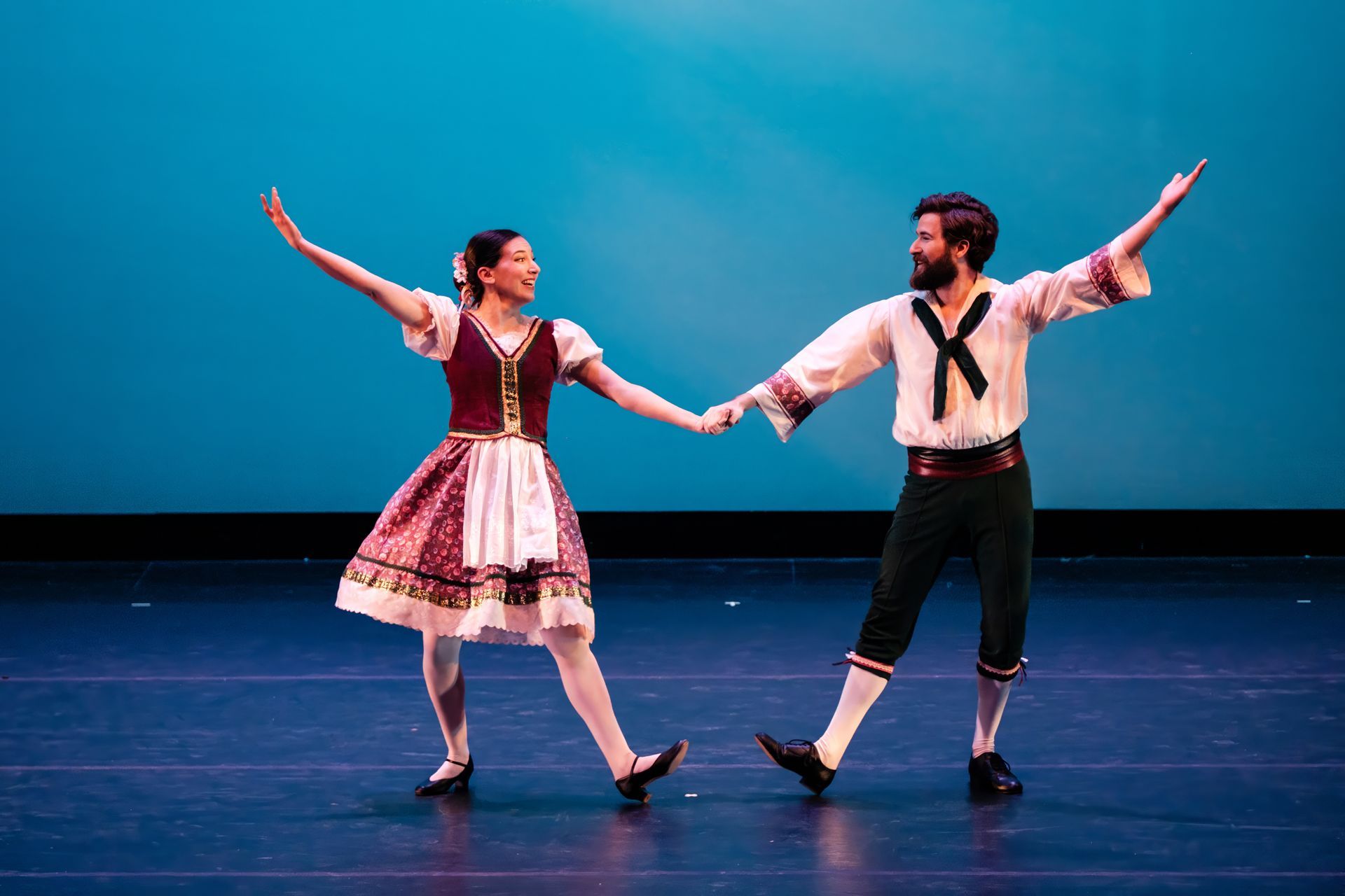 Two dancers in traditional costumes holding hands and posing on a stage with a blue backdrop