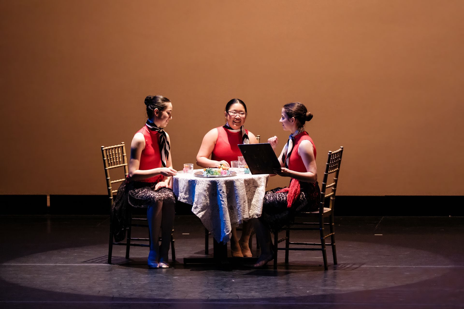 Three performers in red sit around a small table on a dark stage with a plain brown backdrop.