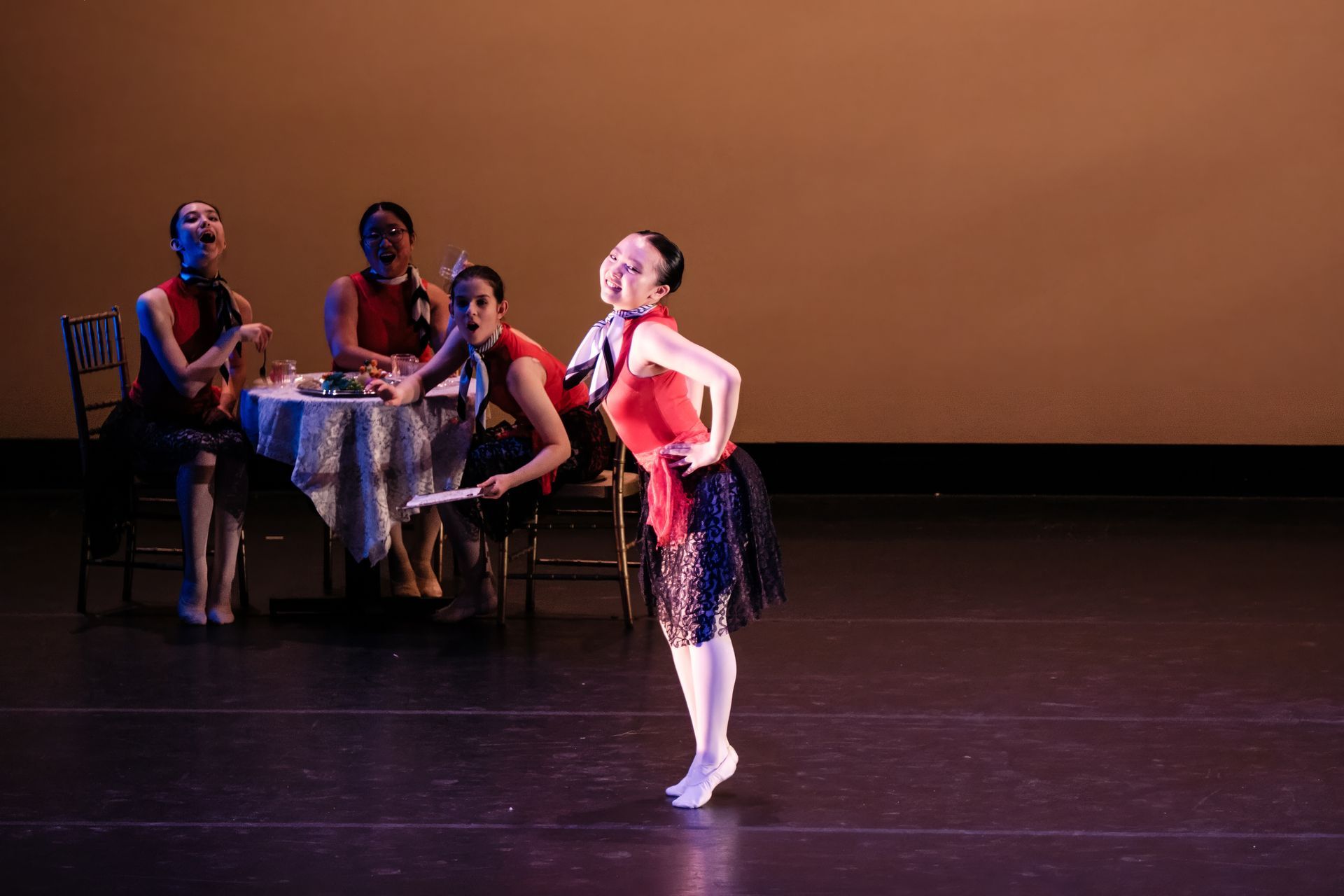 Ballet dancer in red leotard posing on stage beside seated performers and a small table