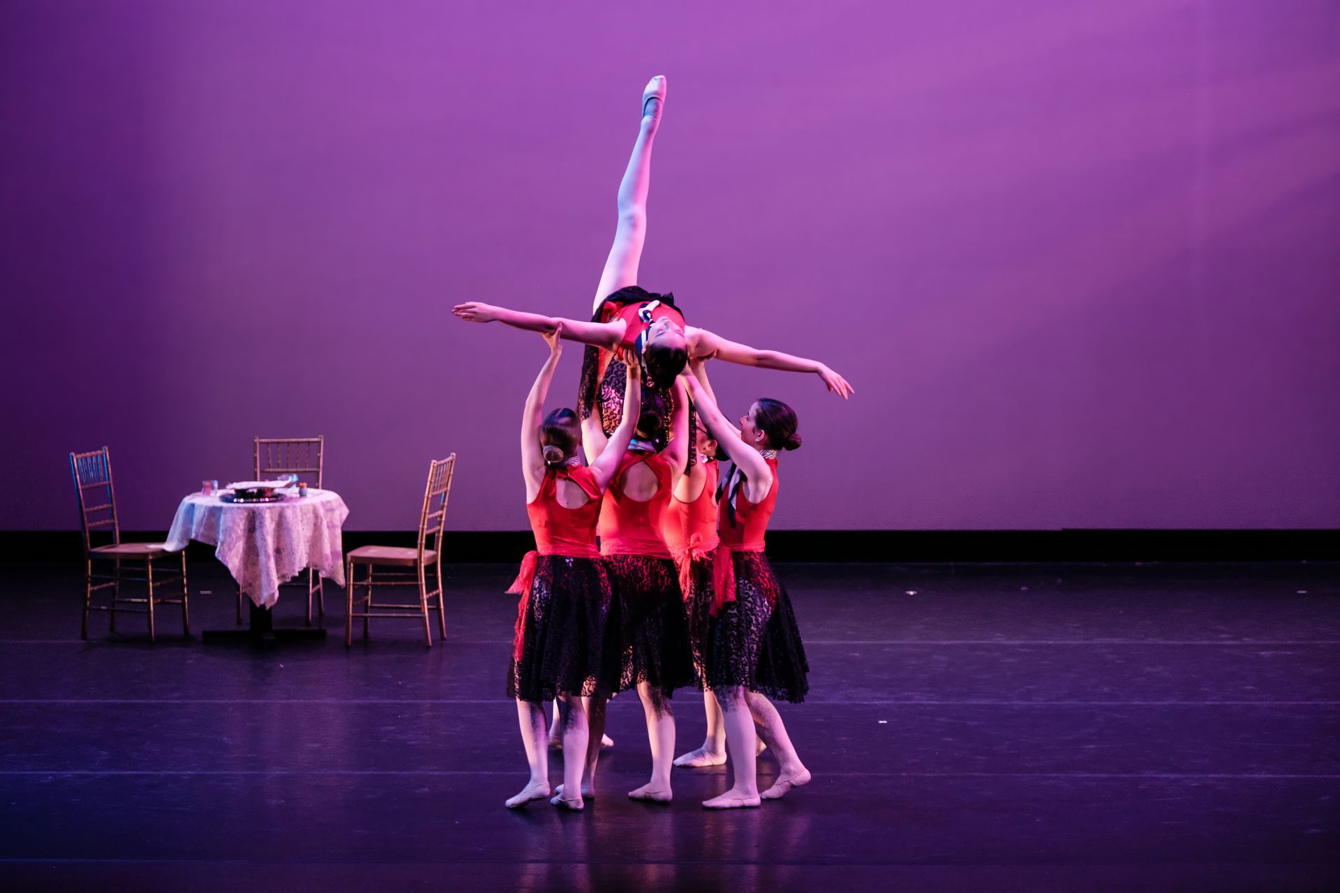 Dancers in pink tops and black skirts form a stacked pose on a purple-lit stage, with a table and chairs at left.