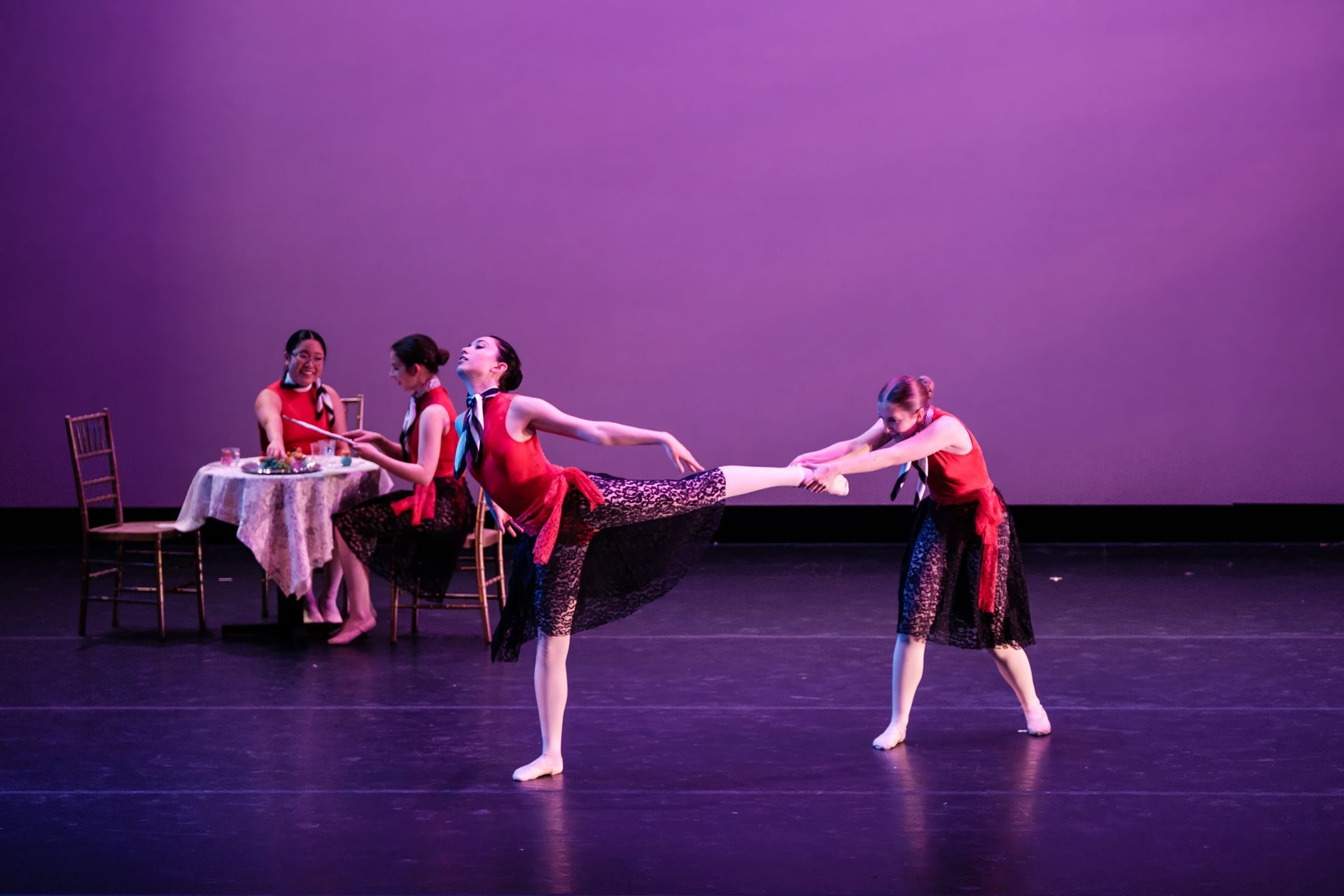 Two dancers in red and black balance in a ballet pose on a purple-lit stage, with a seated table scene behind them