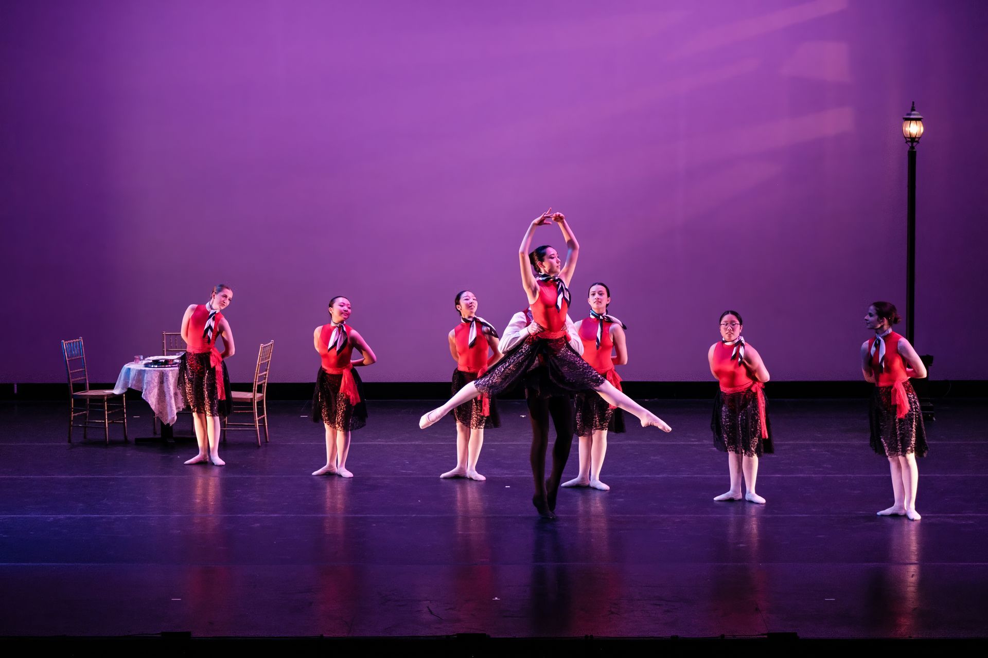 Ballet dancers in red costumes perform on a stage with purple lighting and a piano in the background