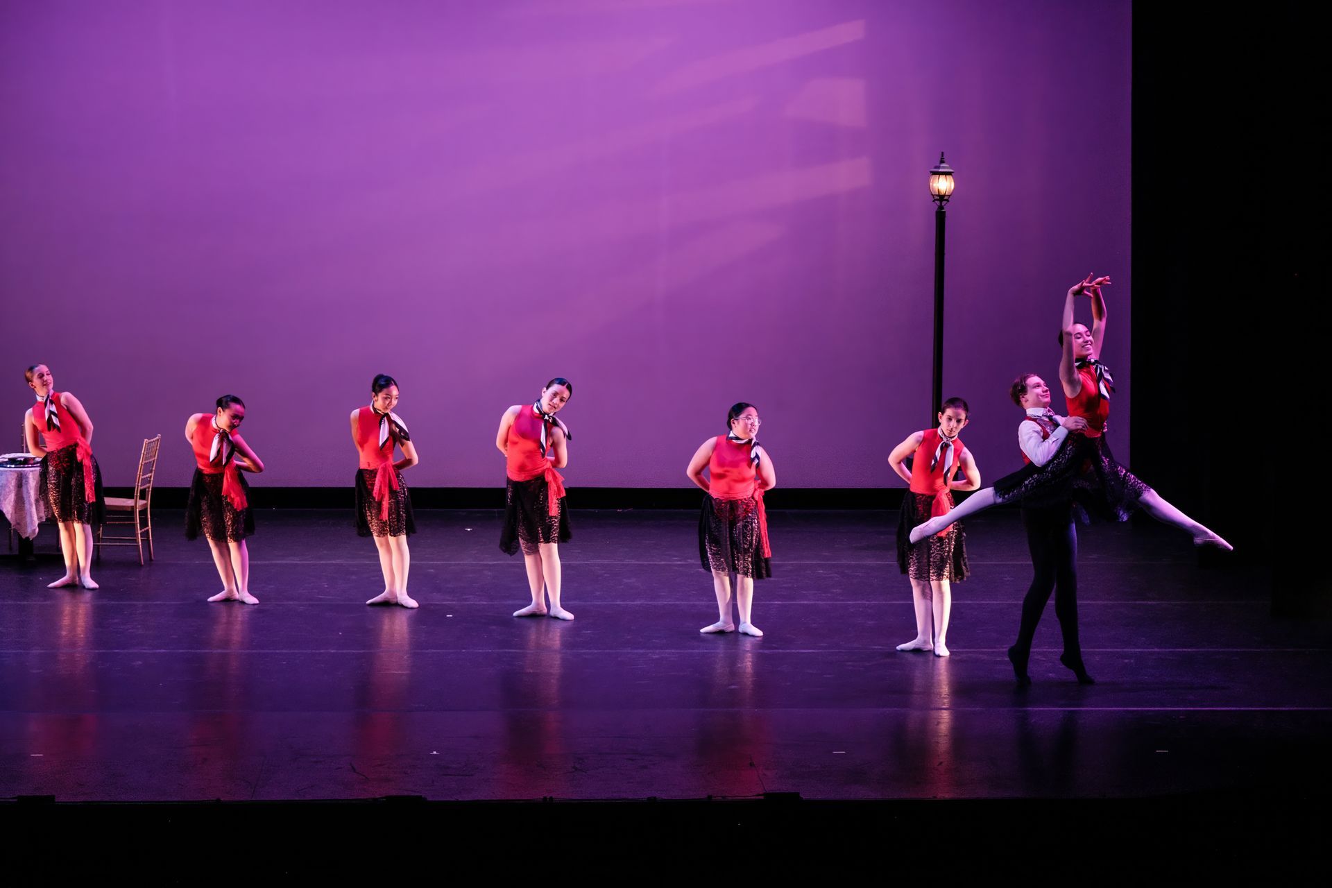 Dancers in red costumes perform on a purple-lit stage with a tall lamp post prop.