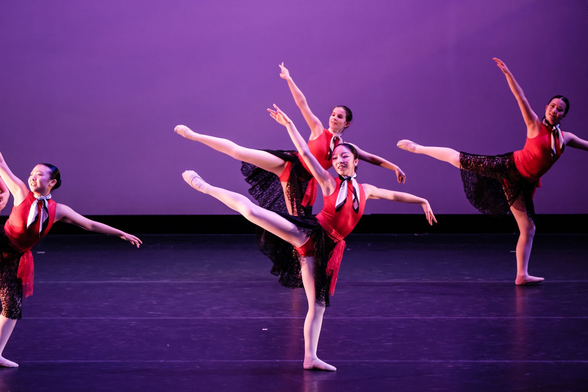 Four dancers in red costumes perform ballet poses on a purple-lit stage