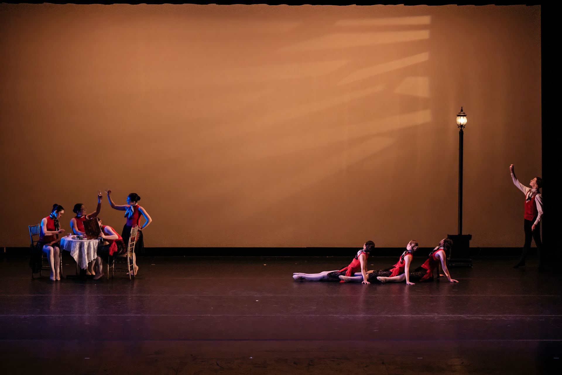 Dancers on a dim stage with a large tan backdrop, some clustered left and others reclining center, near a lit lamp.