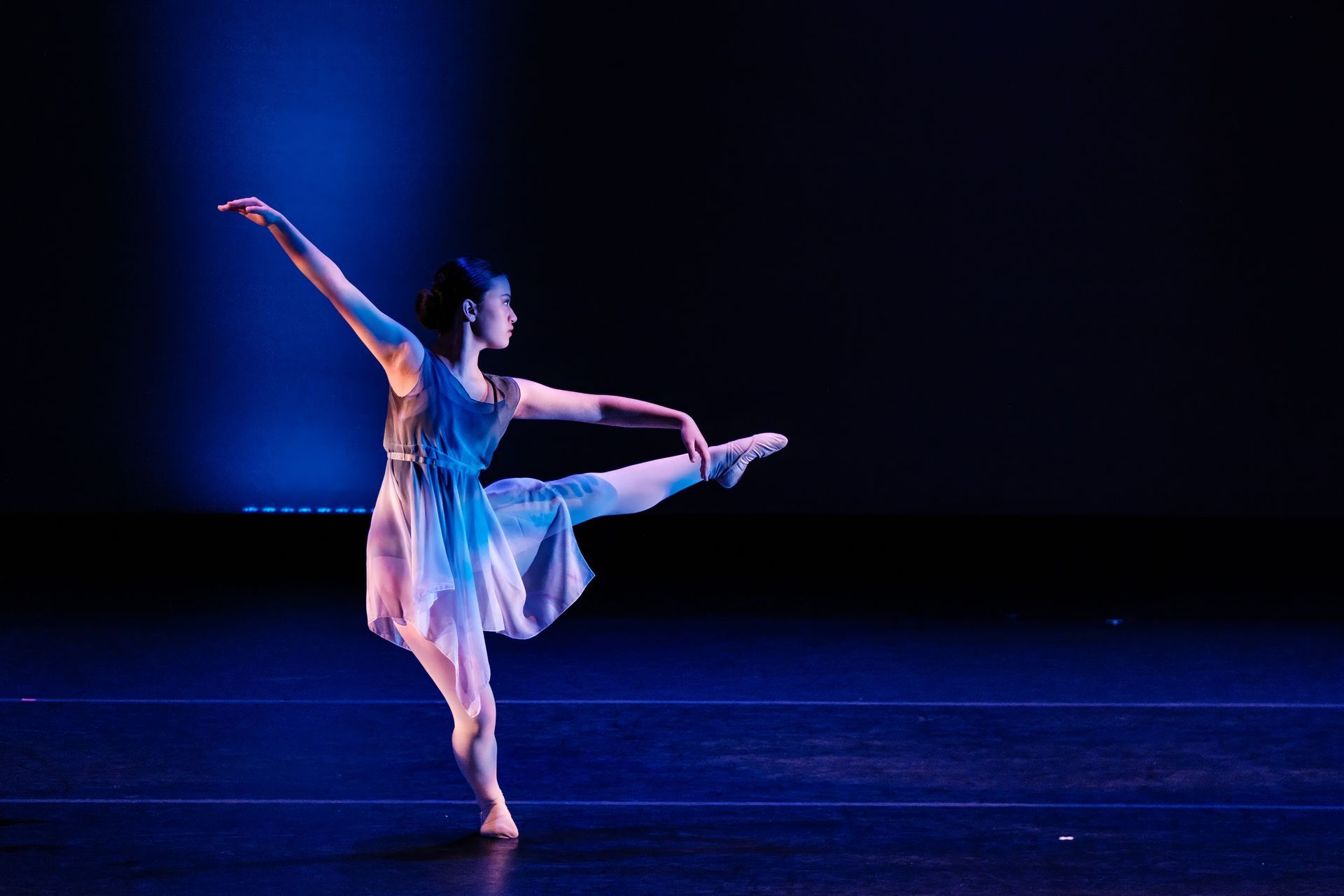 Ballet dancer in a white dress balancing on one leg under blue stage lighting