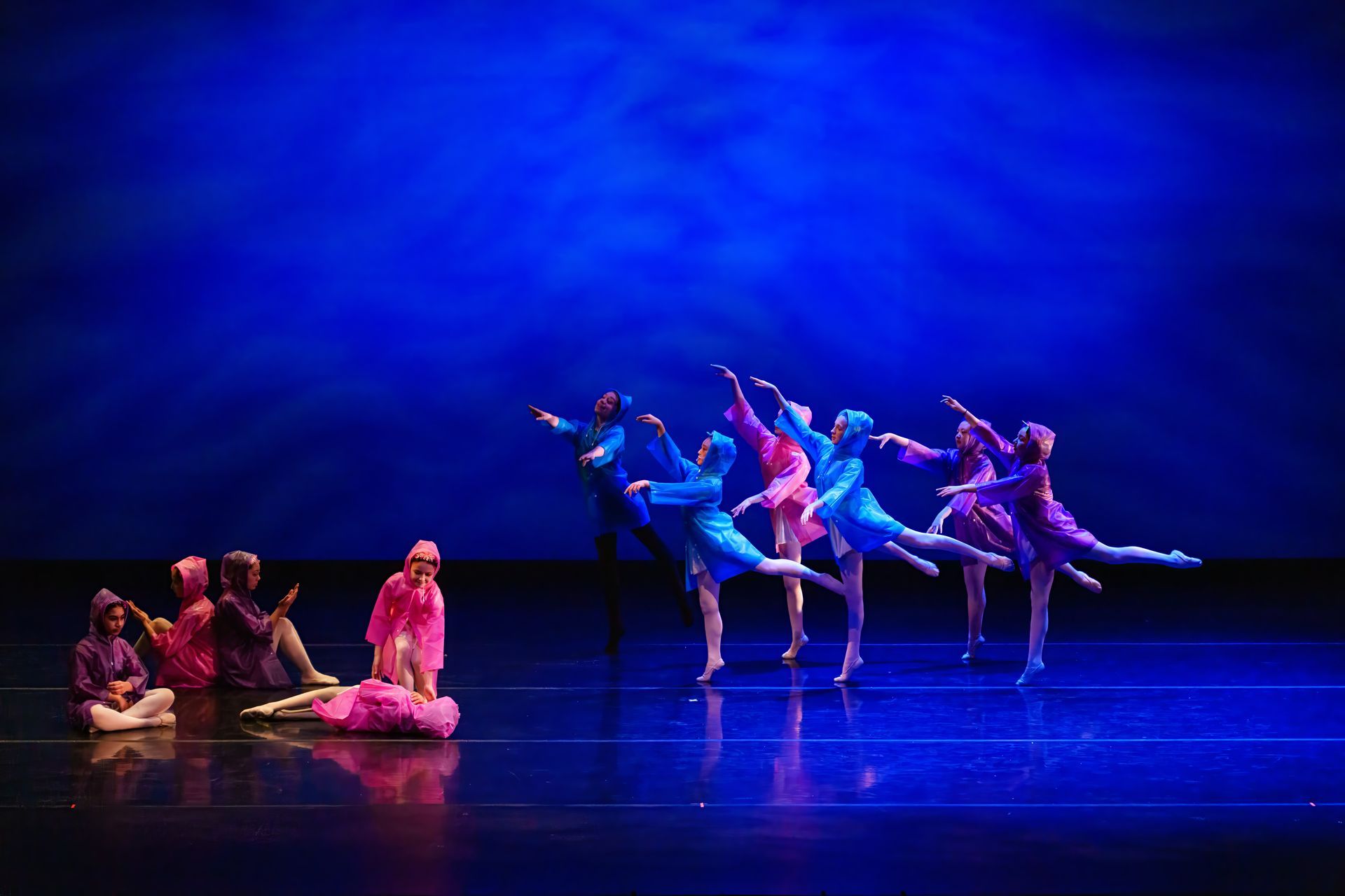 Ballet dancers in colorful costumes performing on a blue-lit stage, with seated dancers at left.
