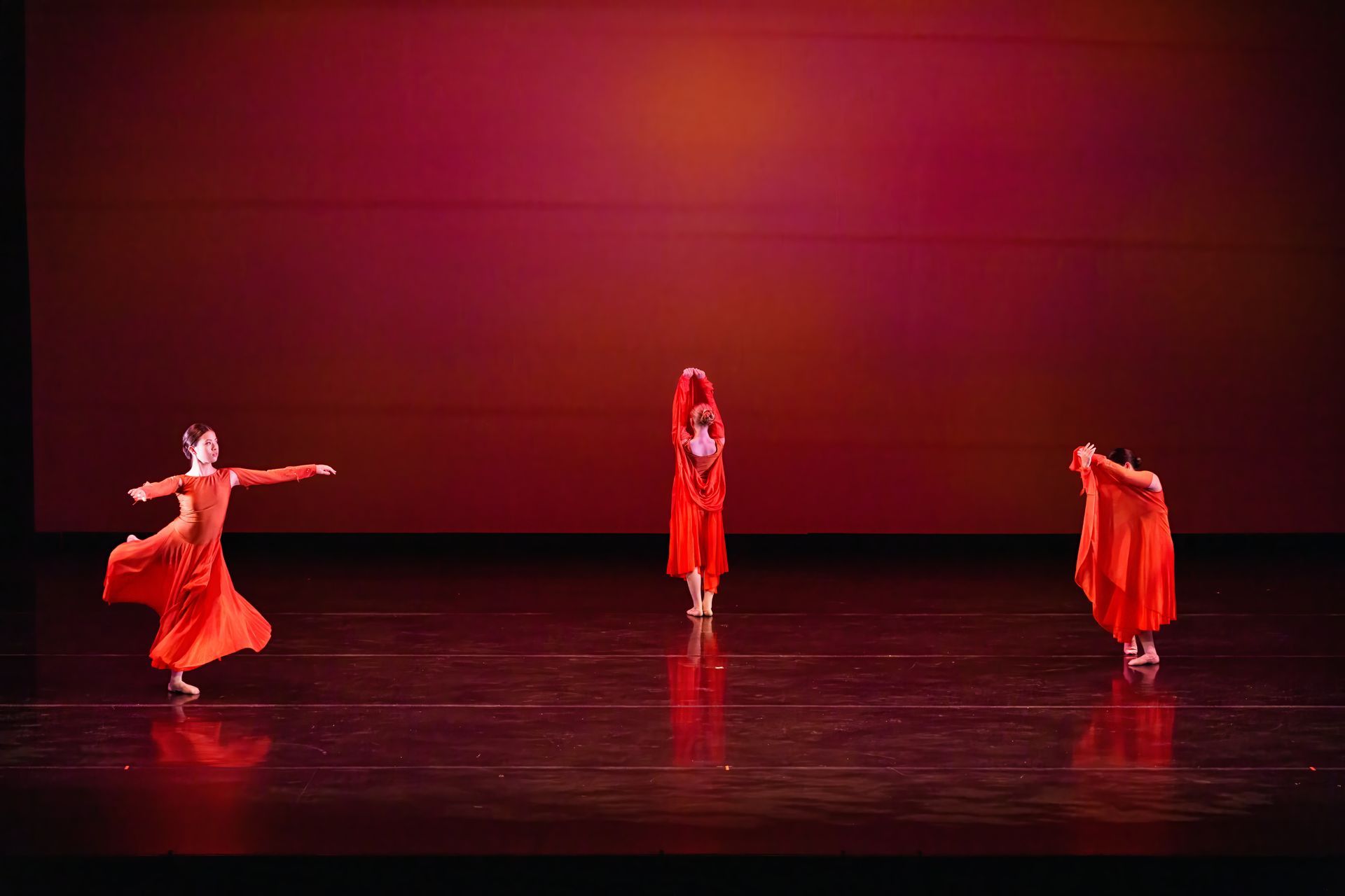 Three dancers in red costumes perform on a dimly lit stage with a red backdrop.