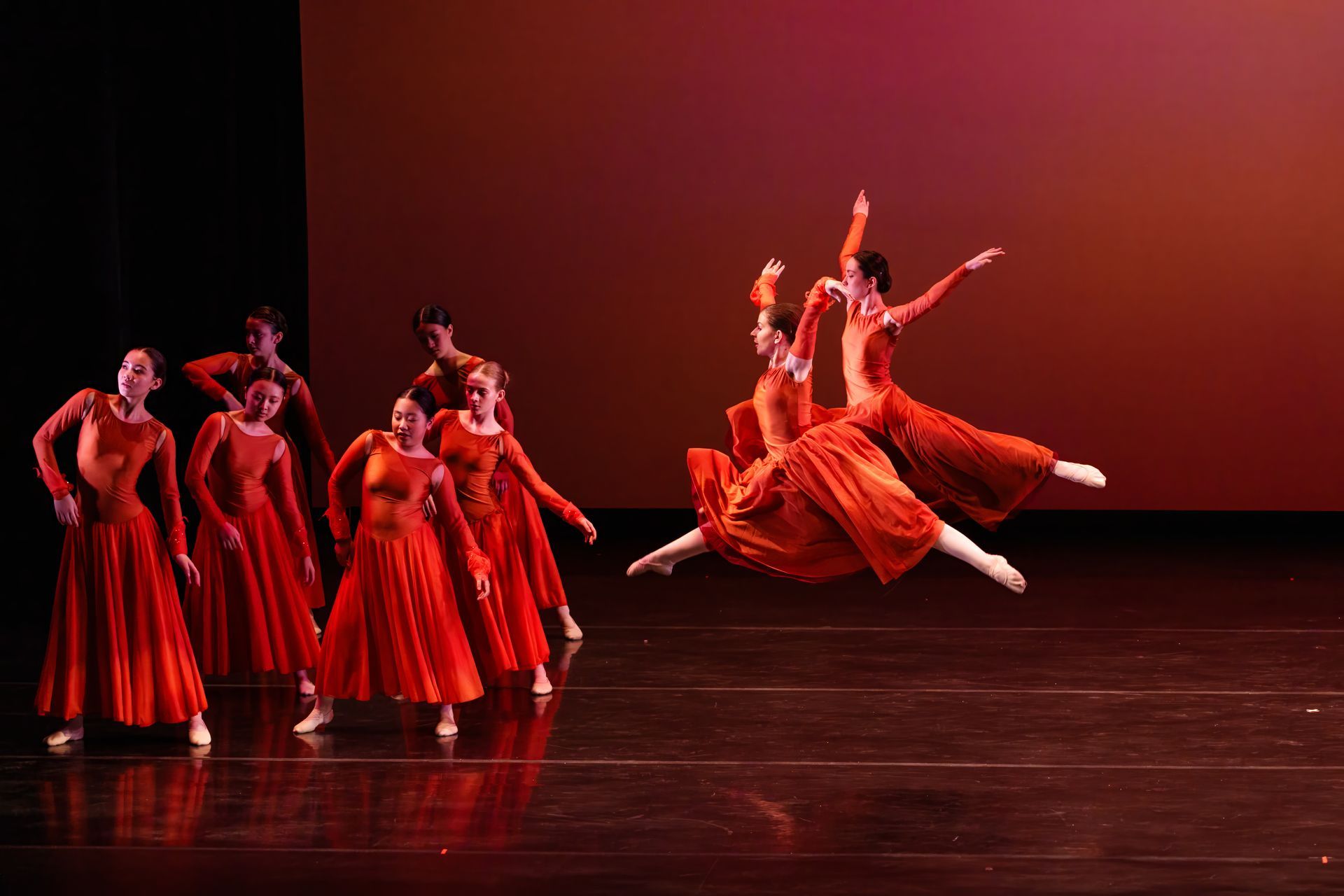 Red-costumed dancers performing a ballet leap on a stage under warm lighting