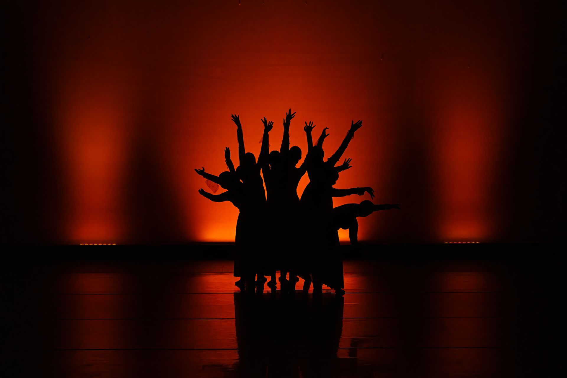 Silhouetted dancers with outstretched arms on a red-lit stage reflected on a glossy floor