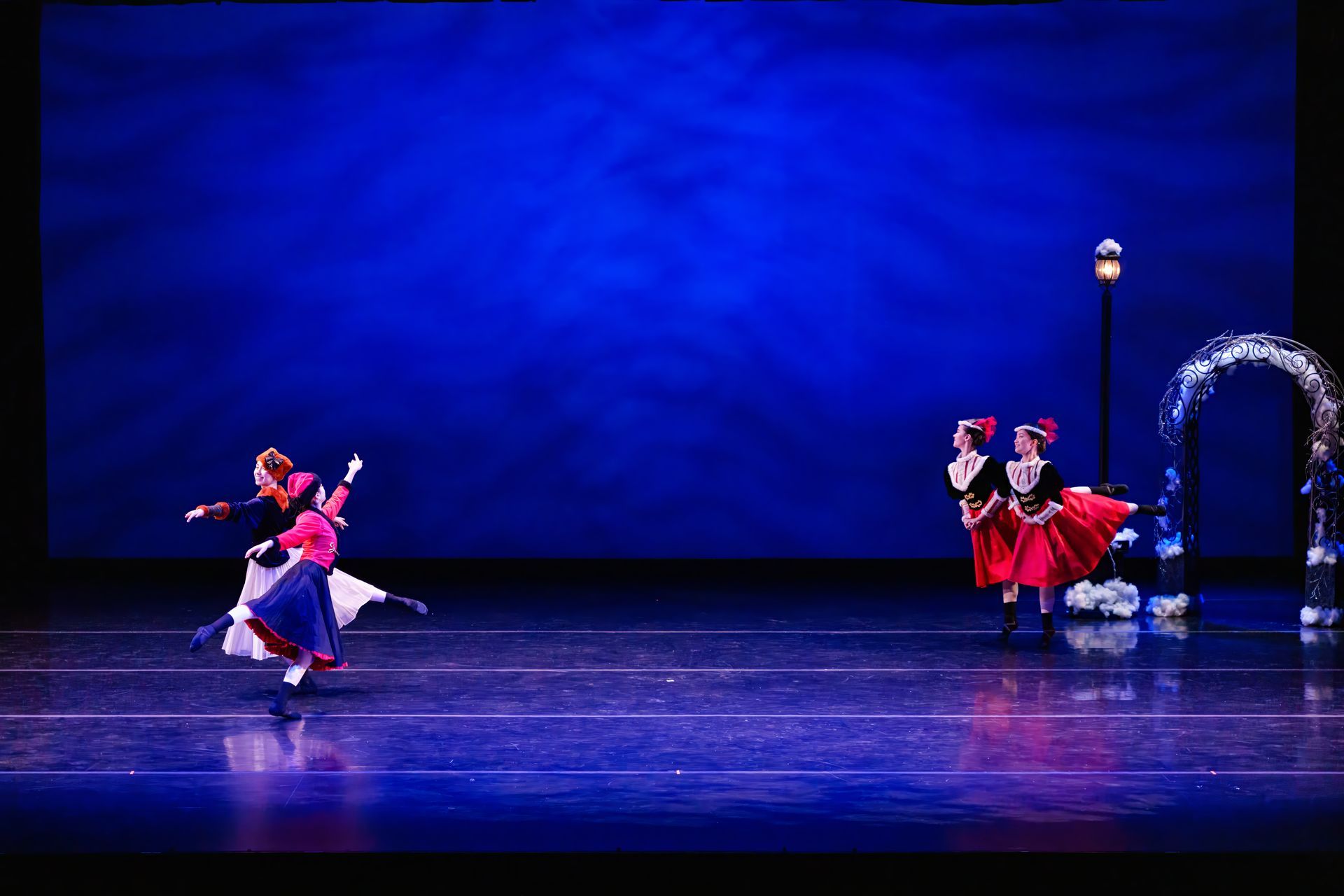 Two dancers in red and white costumes perform on a blue-lit stage near a decorated arch.