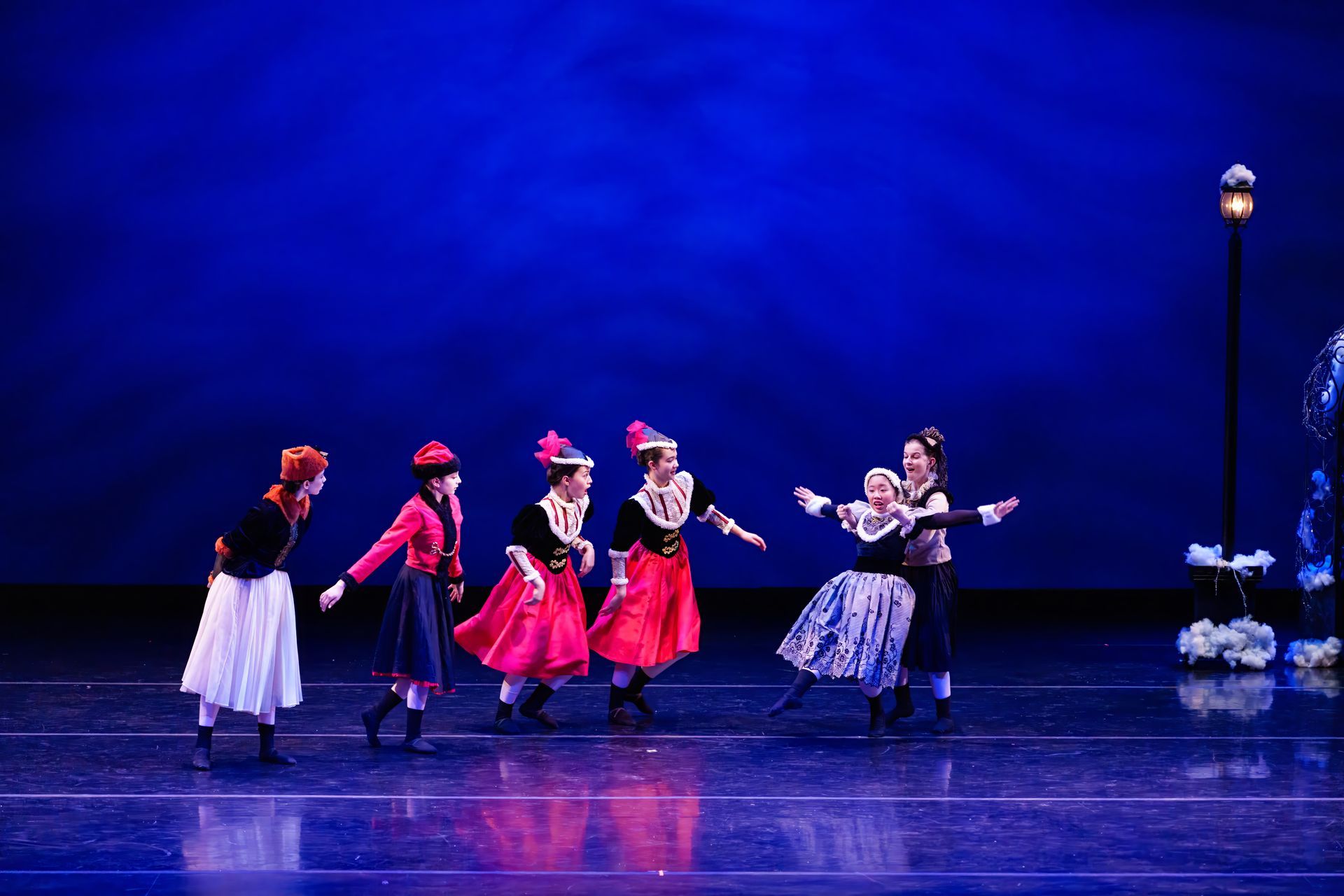 Five dancers in colorful costumes perform on a blue-lit stage near a streetlamp prop.