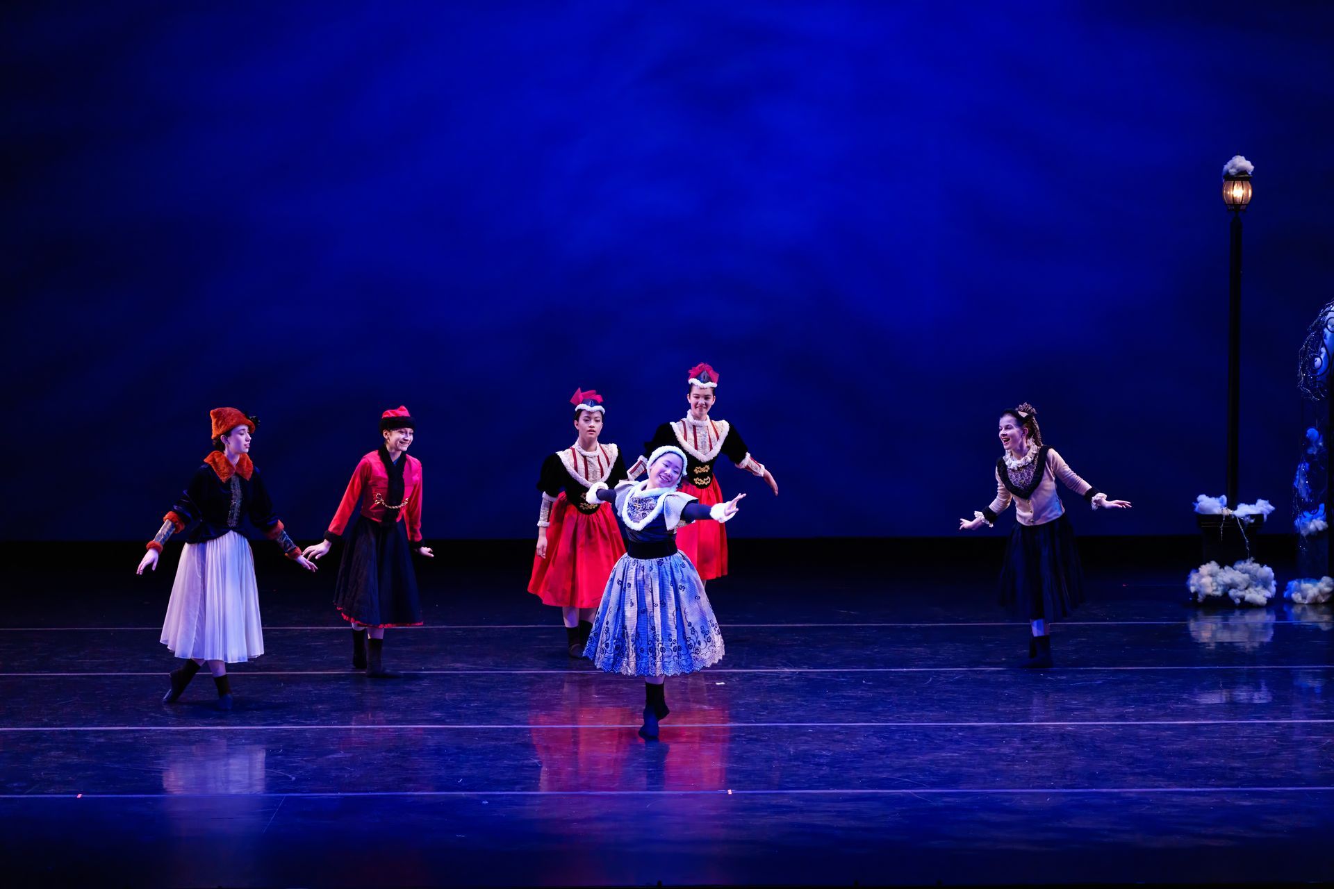 Five dancers in colorful folk costumes perform on a blue-lit stage with a lamppost backdrop