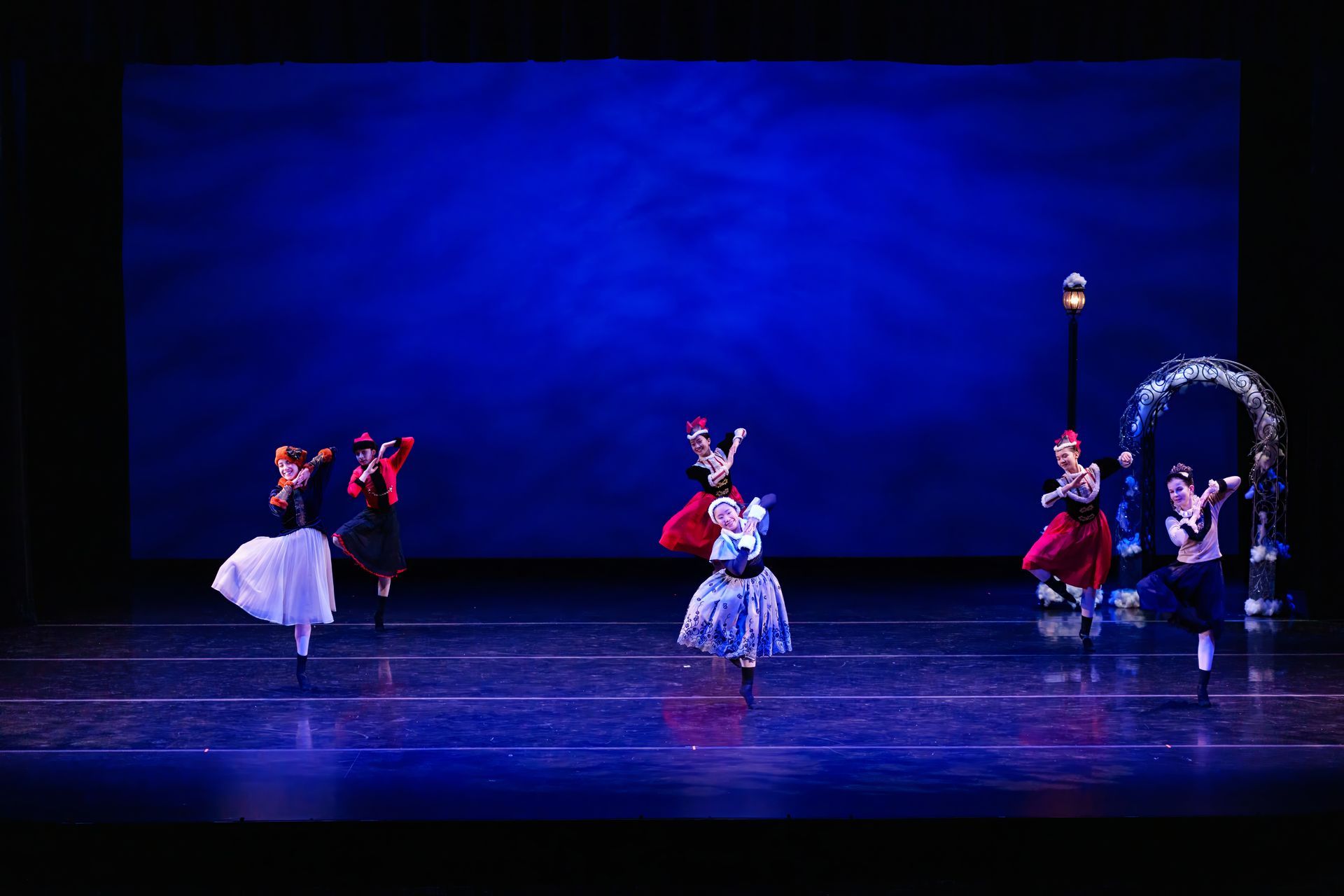 Ballet dancers in colorful costumes performing on a blue-lit stage with a circular prop