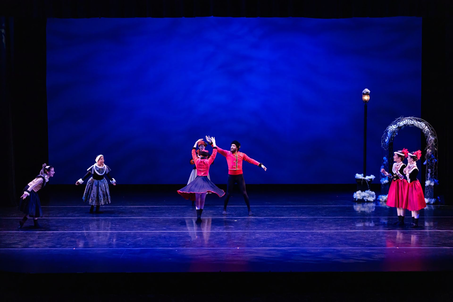 Dancers in red and black costumes perform on a blue-lit stage with a lamppost and archway set.