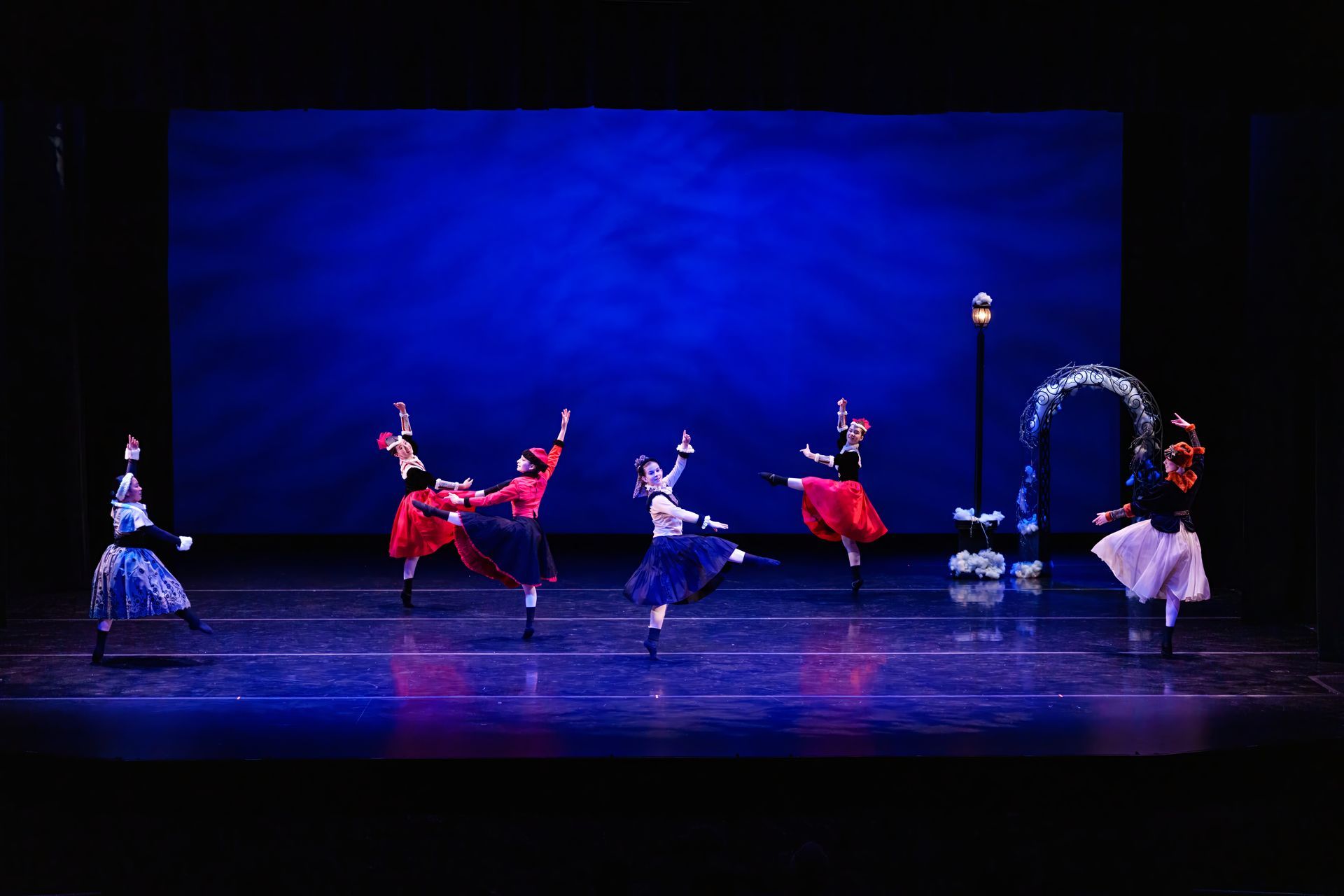 Ballet dancers in red and blue costumes perform on a stage with a blue backdrop and arch set piece