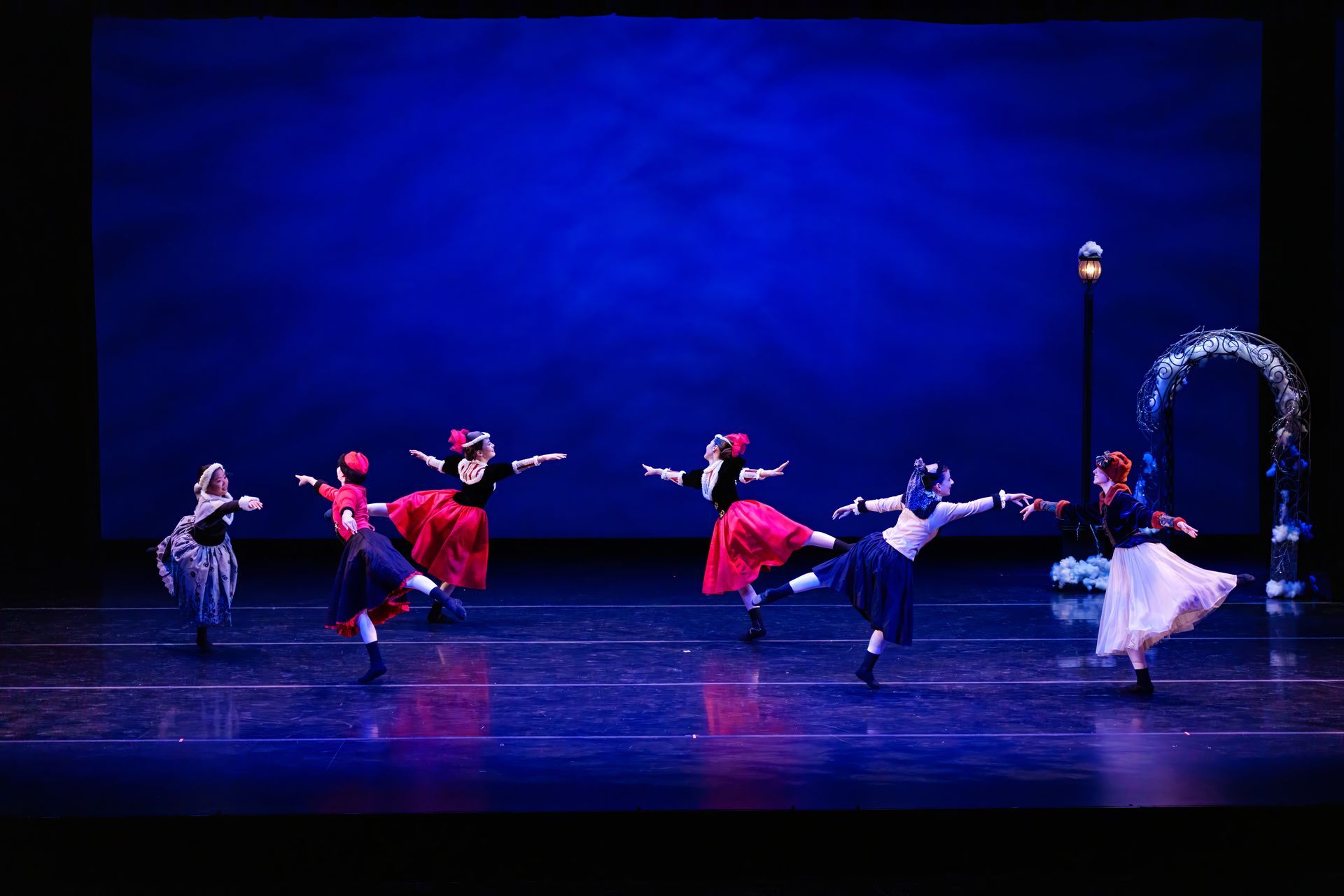 Dancers in colorful costumes perform on a blue-lit stage, with one dancer posed near a lamp post and arch.