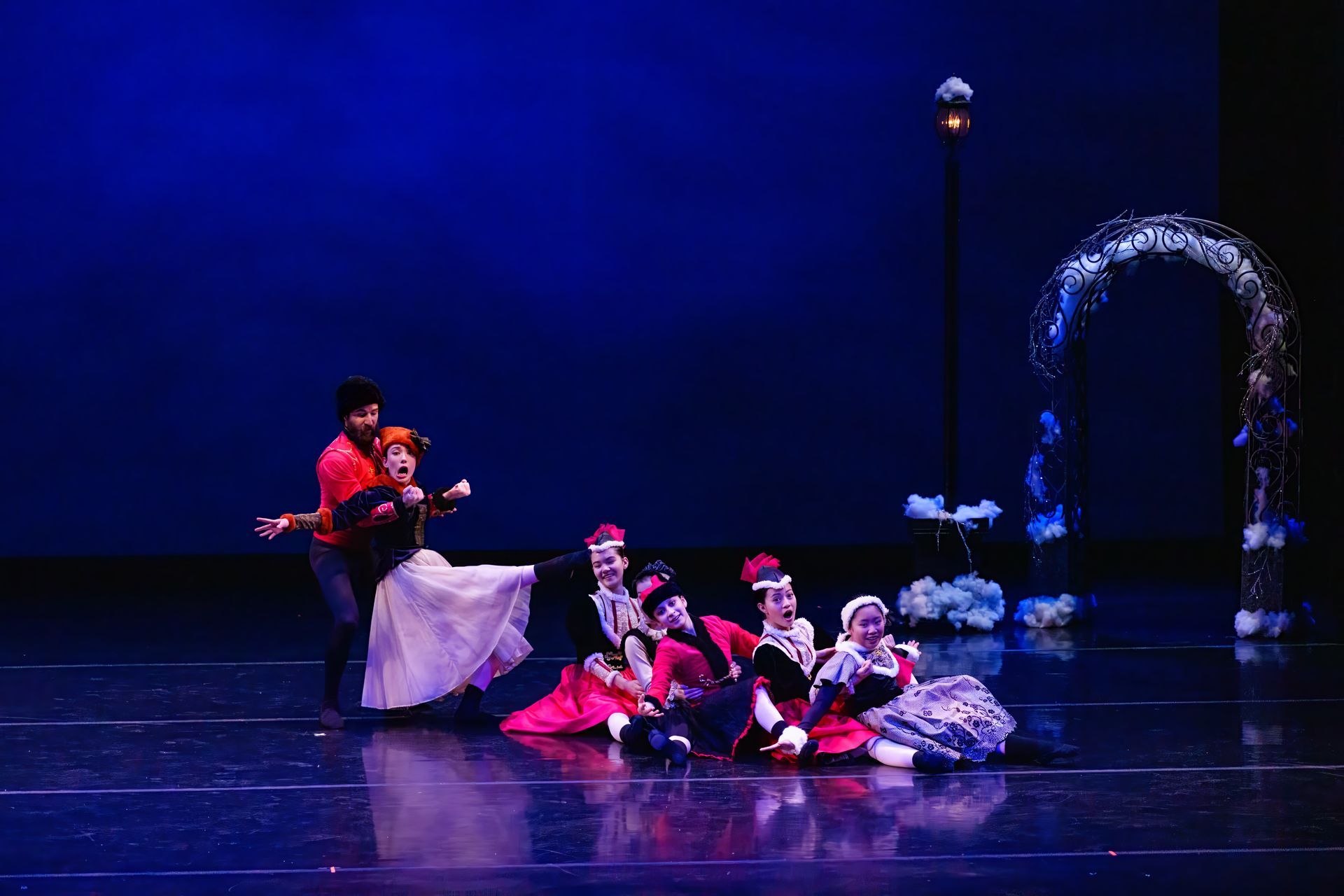 Stage performers in red and white costumes sit on a dark stage beside a glowing fountain arch.