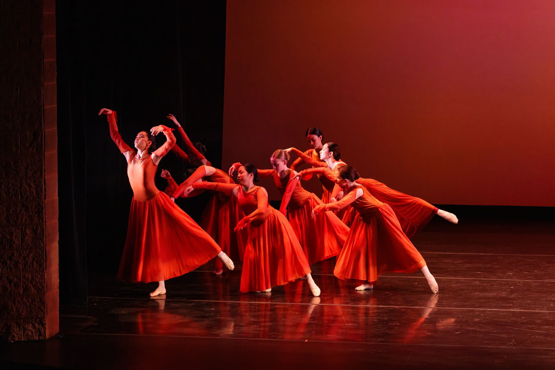 Dancers in flowing red costumes perform a ballet on a stage with warm red lighting.