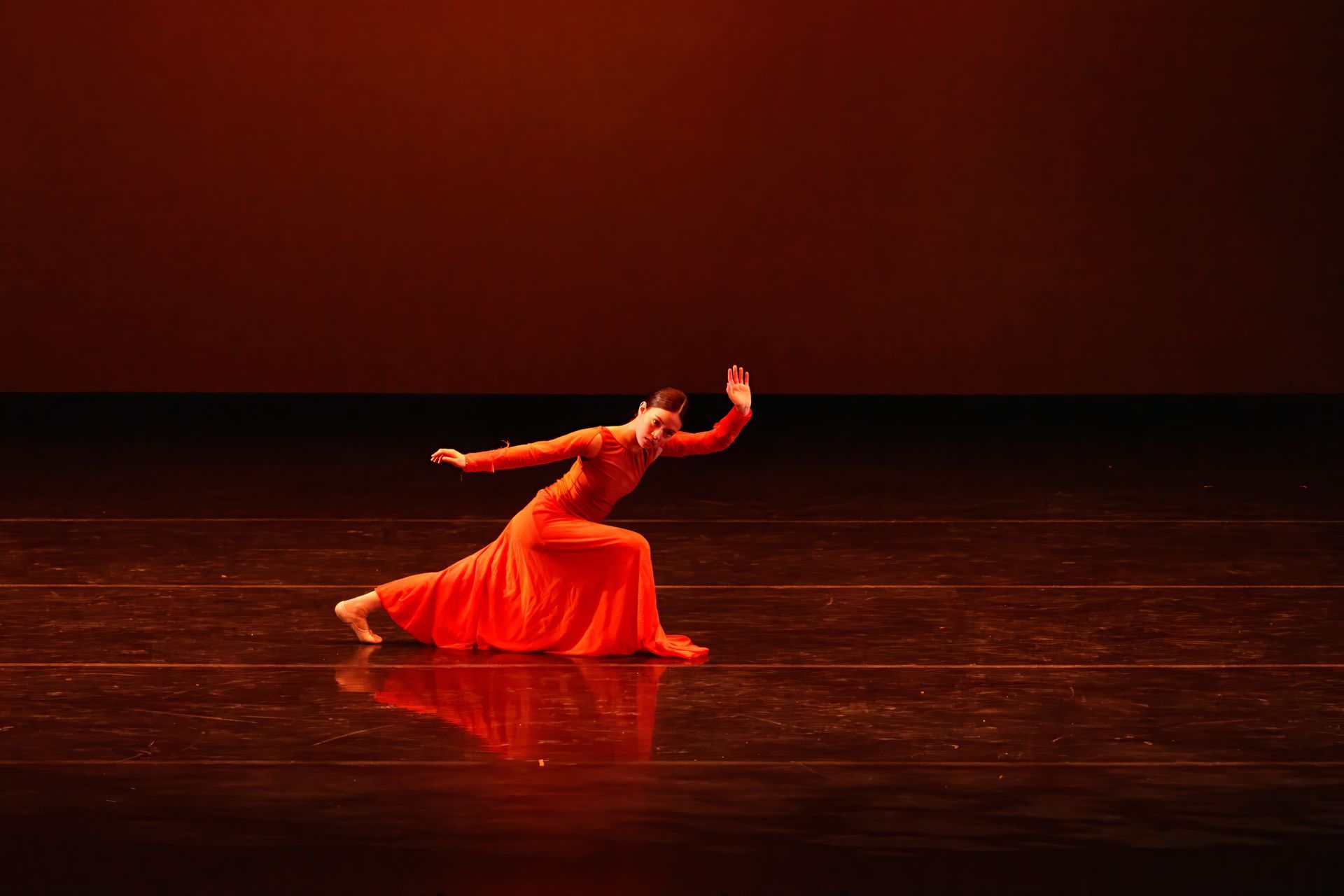 Dancer in flowing red costume kneels and reaches upward on a dark stage with warm red lighting