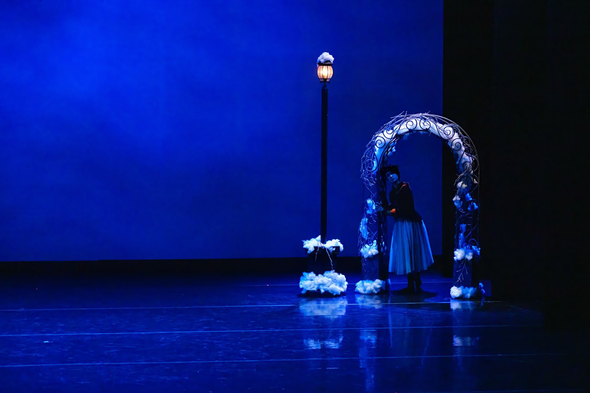 Blue-lit stage with a person standing beside a glowing arch and a tall lamp-like post