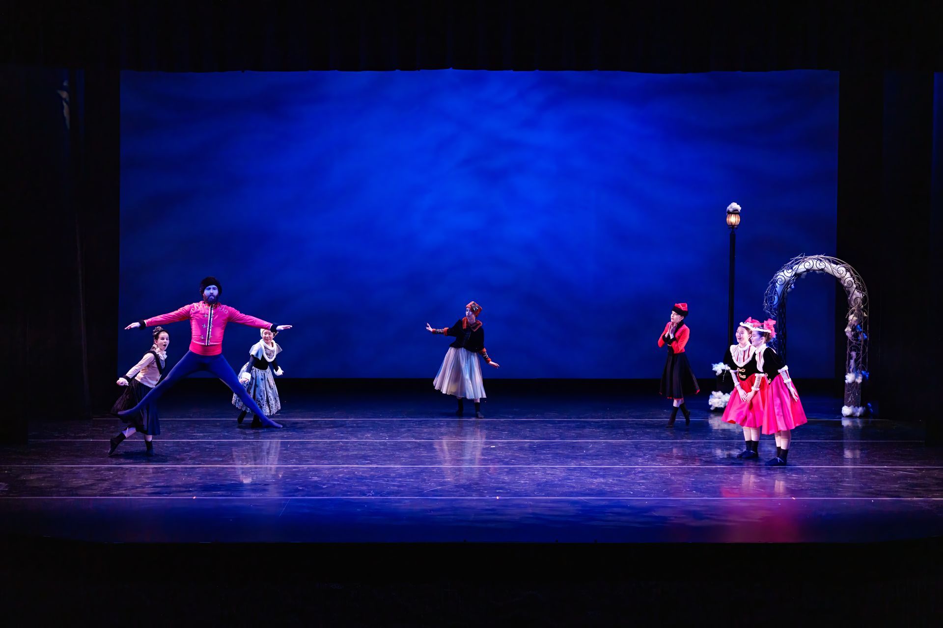 Dancers in colorful costumes perform on a blue-lit stage with a lamppost and arch backdrop.