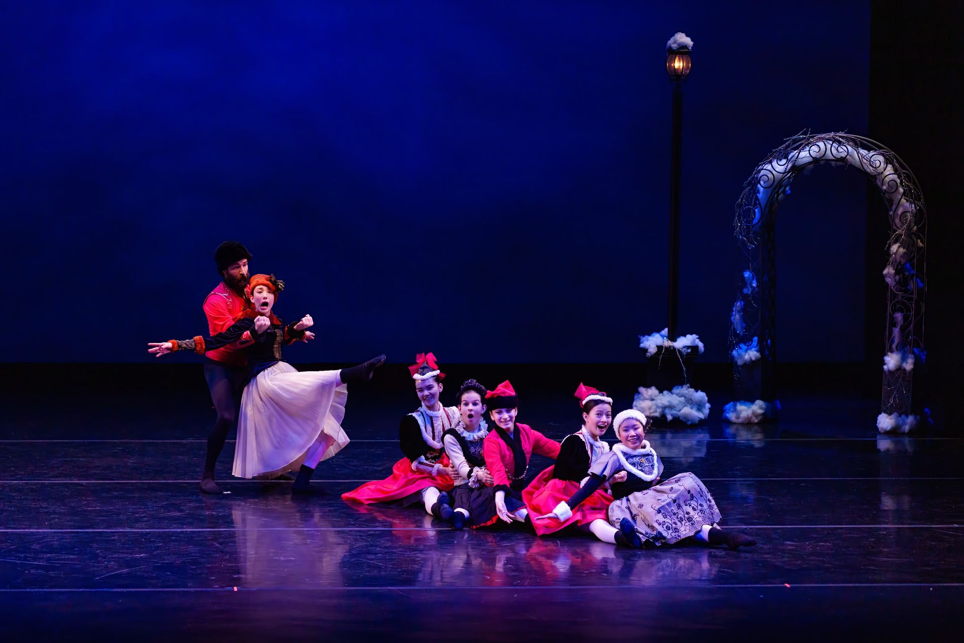 Costumed dancers perform on a dark stage with blue lighting and a decorative arch backdrop