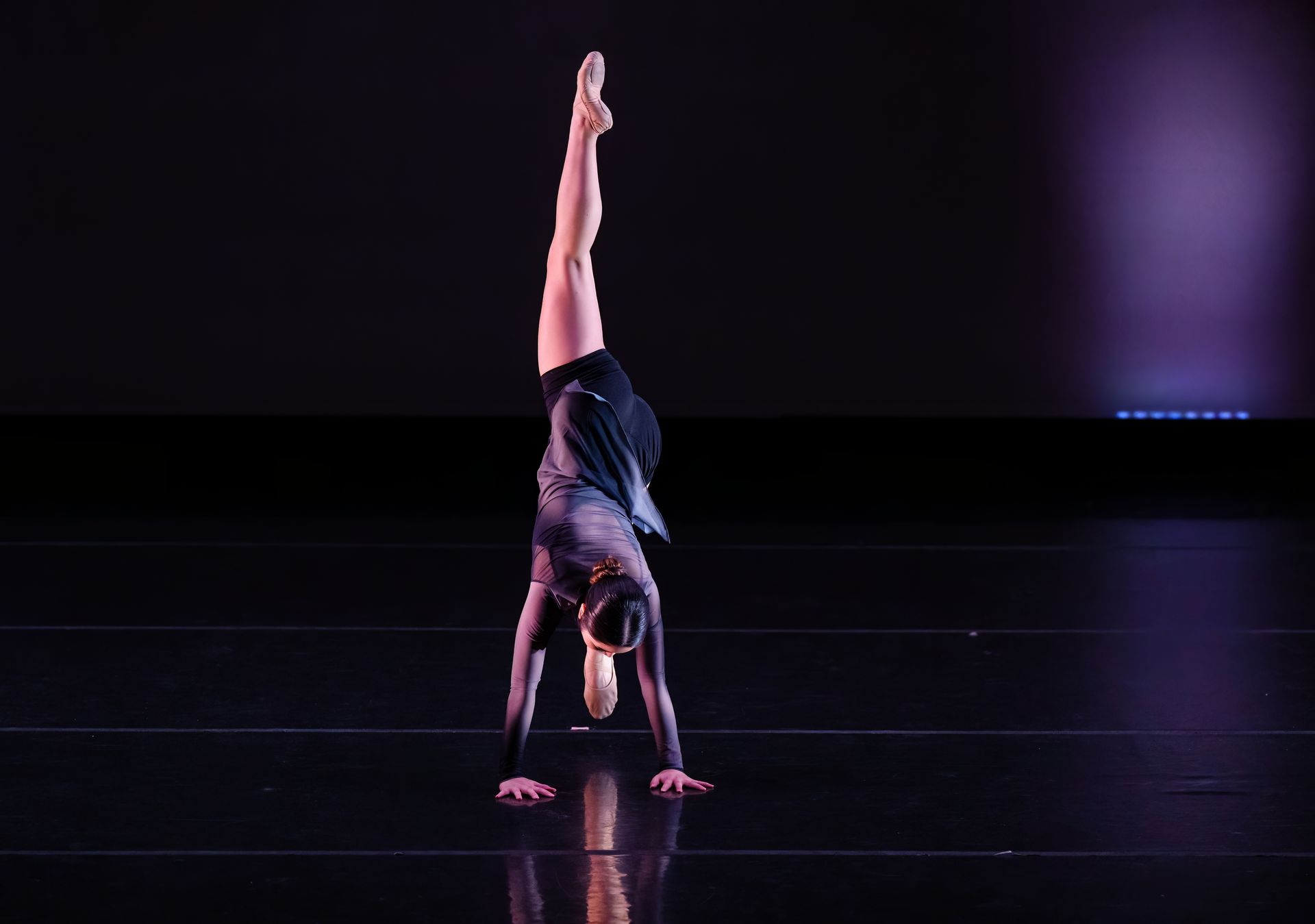 Gymnast doing a handstand on a dark stage, one leg extended straight up under purple lighting