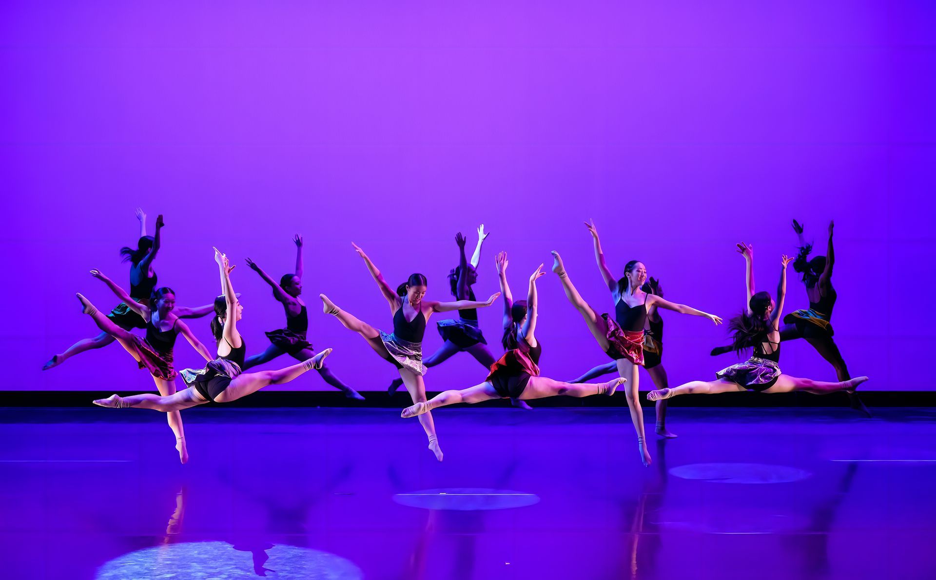 Dancers in black and pink costumes perform synchronized leaps on a purple-lit stage