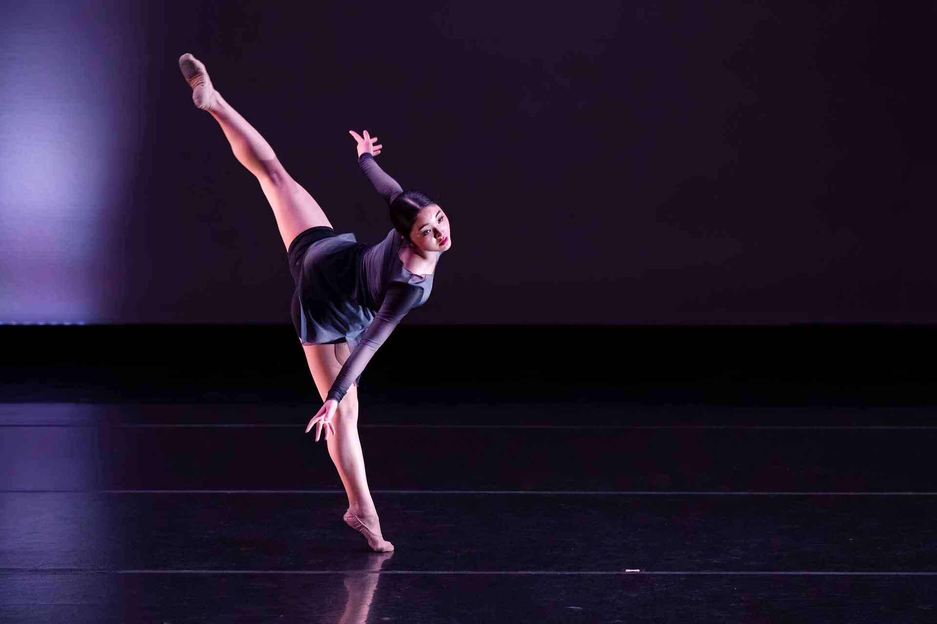 Dancer balancing in a high leg extension on a dark stage with purple lighting