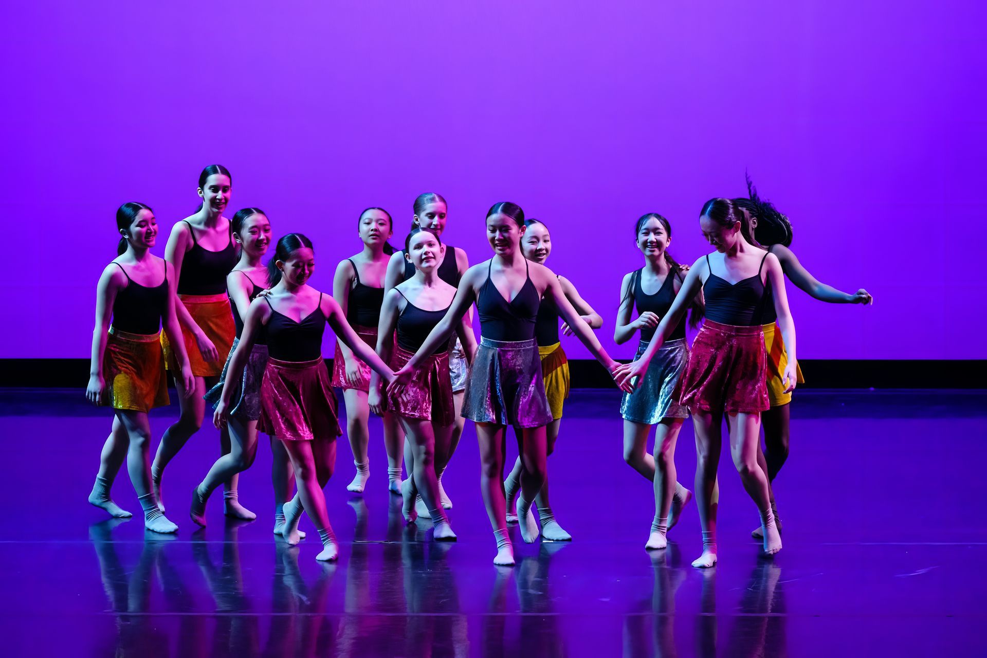 Dance troupe performing on a purple-lit stage, wearing colorful skirts and black tops.