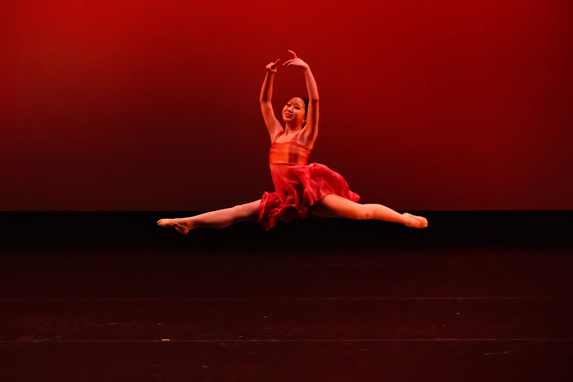 Ballet dancer in red costume performing a center split on a dark stage under red lighting