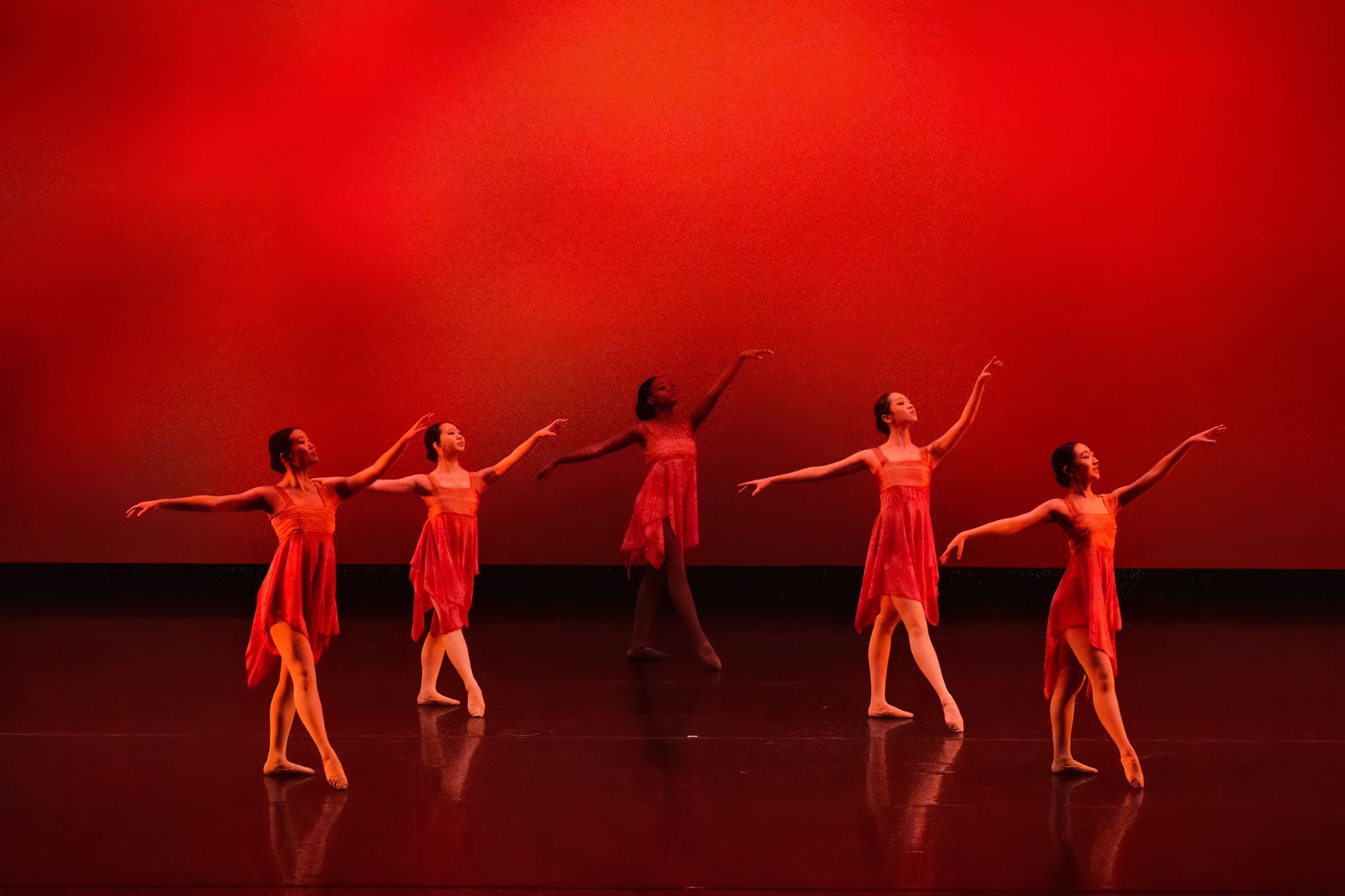 Five dancers in red costumes perform on a stage under red lighting