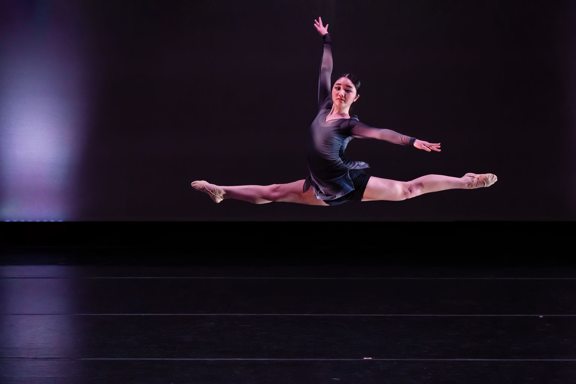 Ballet dancer mid-air in a split leap on a dark stage, wearing a blue costume.