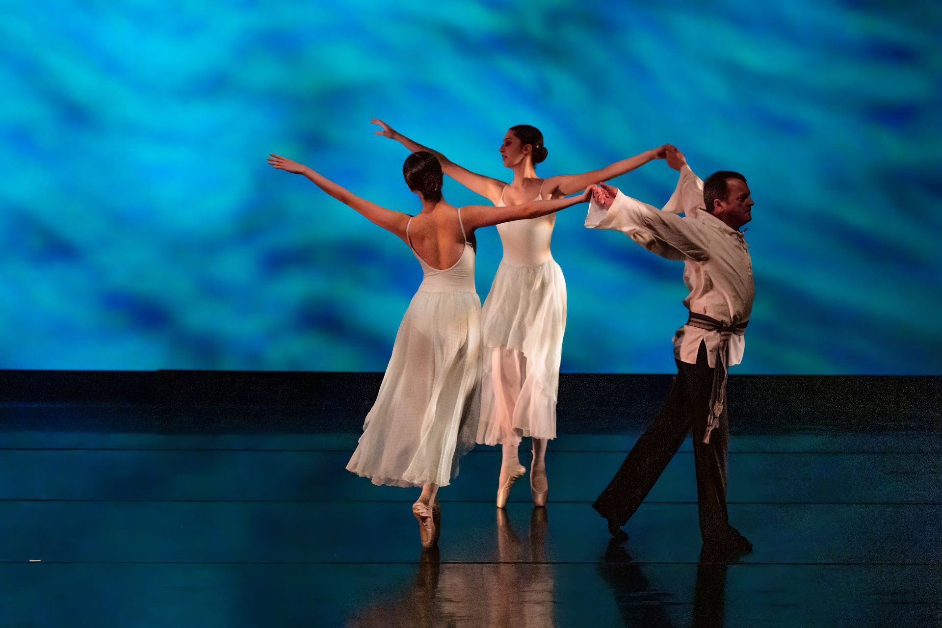 Three dancers in white costumes performing on a stage with a blue backdrop.