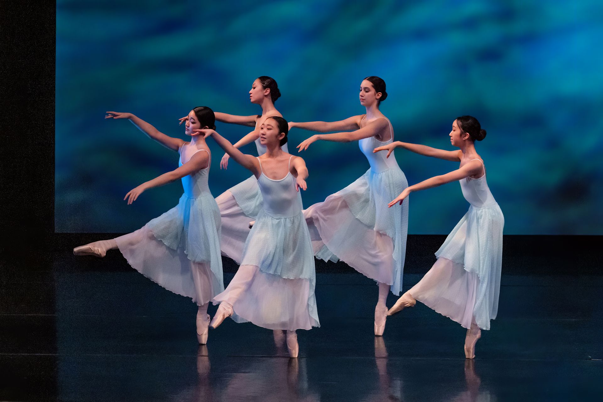 Five ballet dancers in white dresses pose mid-performance on a dark stage