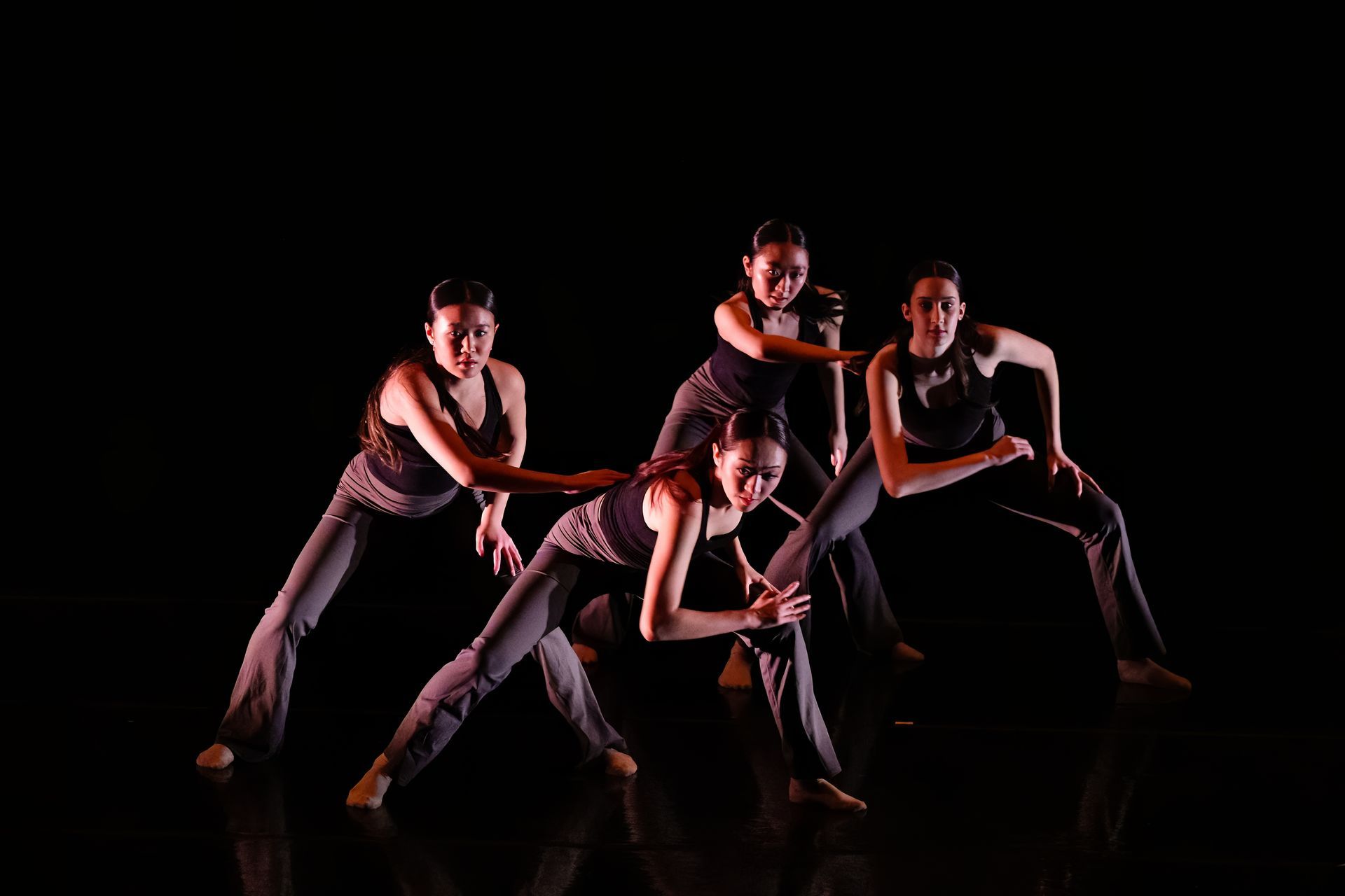 Four dancers in dark outfits pose in a dramatic low-lit studio against a black background.